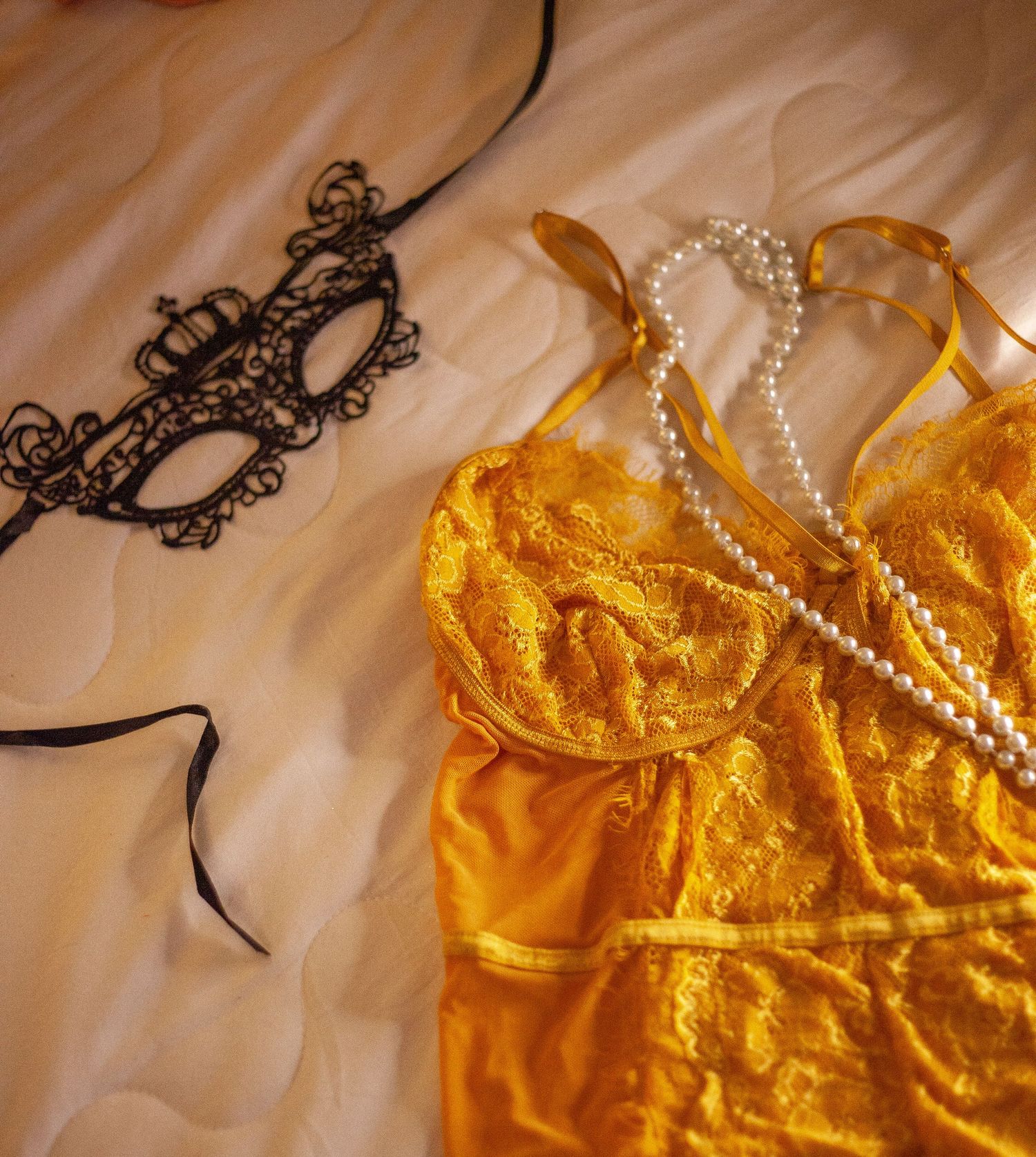 Yellow lace lingerie with pearl necklace and black masquerade mask on silk bedding.