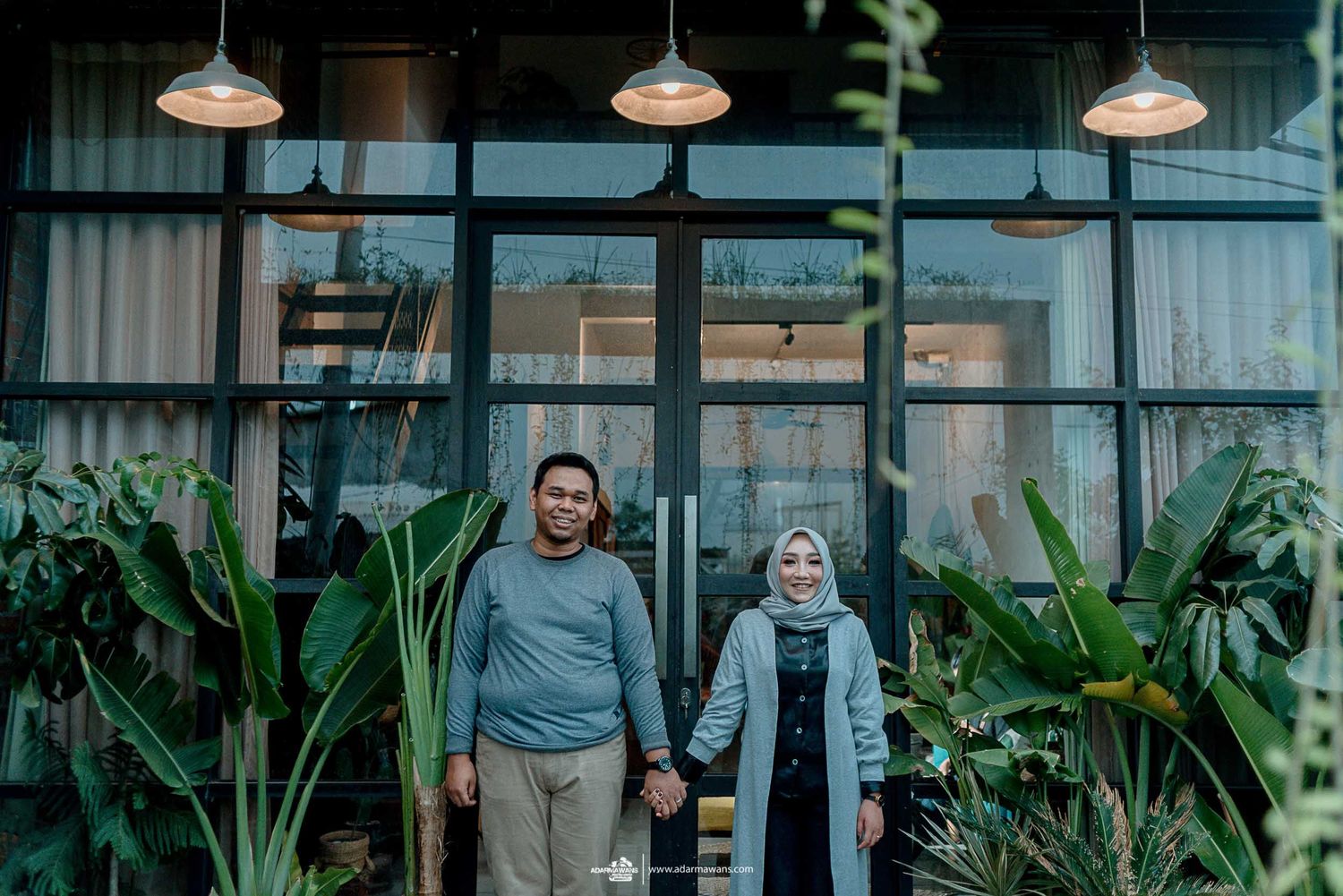 Couple holding hands while standing in front of a modern building with tropical plants and pendant lights.