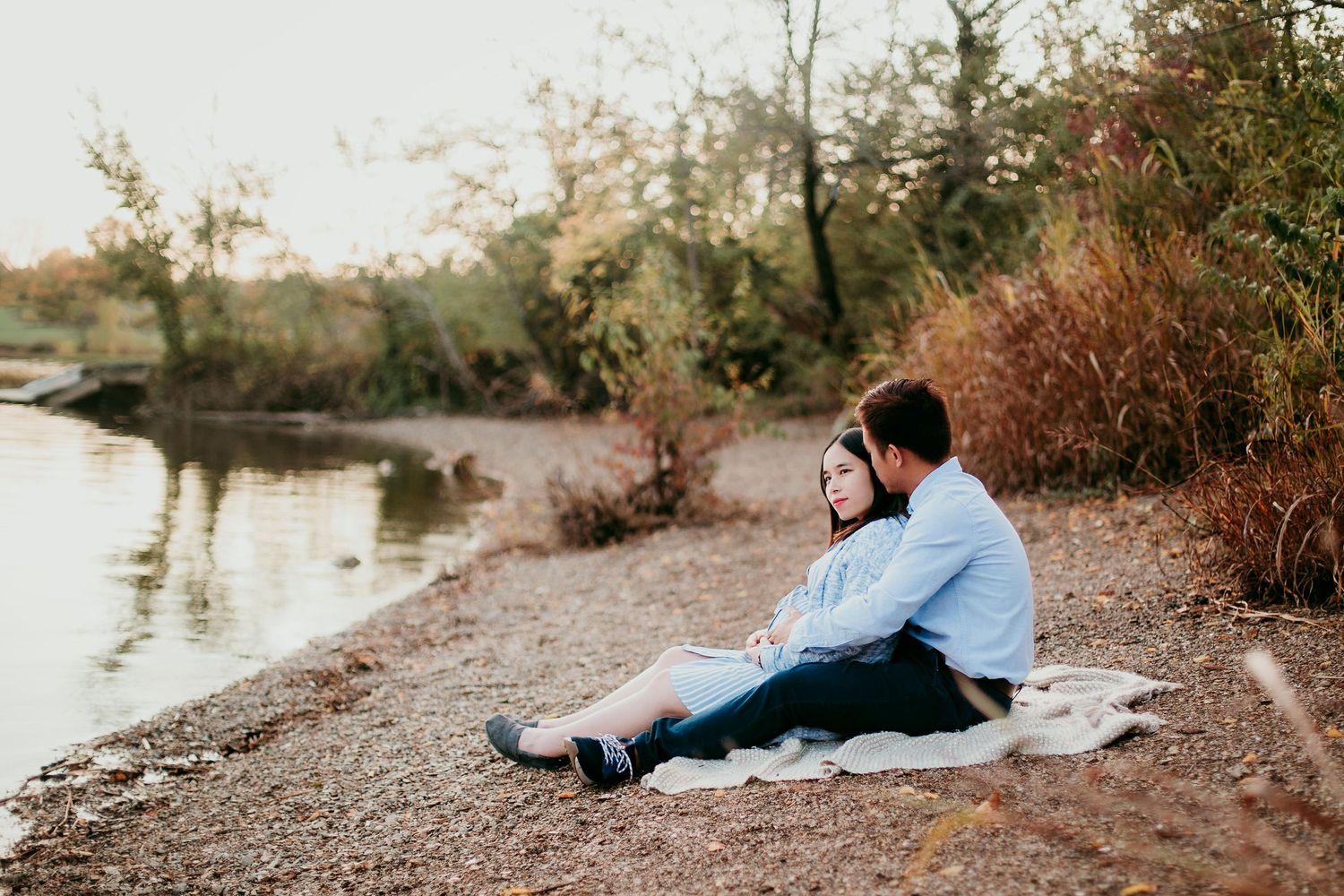 pregnant woman sitting with man on blanket at edge of waters edge at longview lake in lee's summit