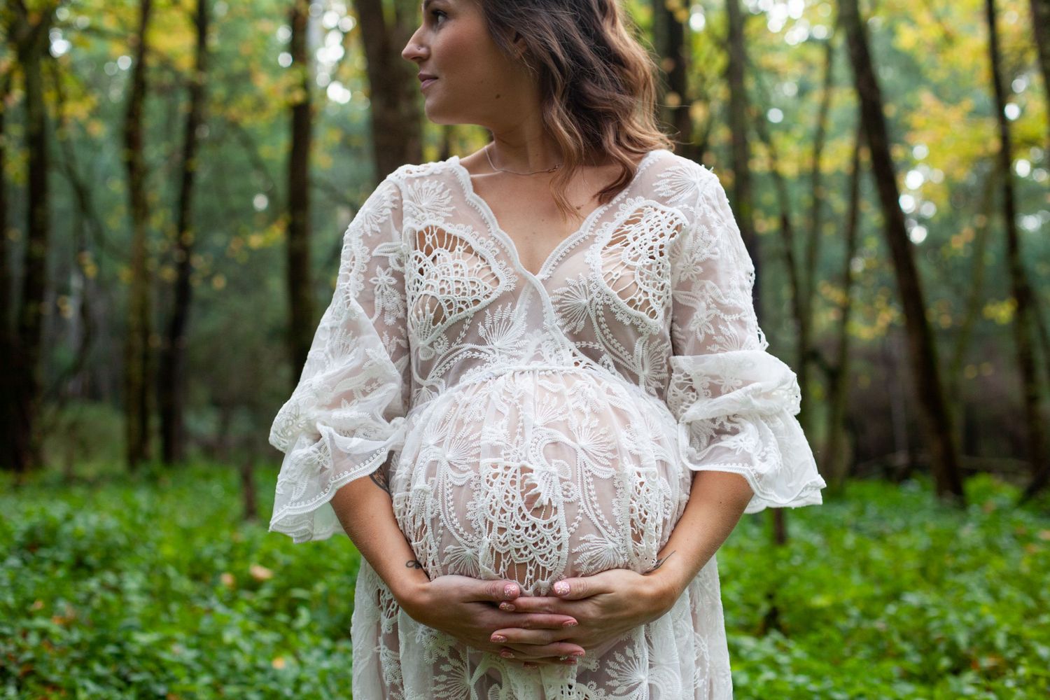 Ethereal maternity portrait featuring a white lace dress in a lush green forest setting.