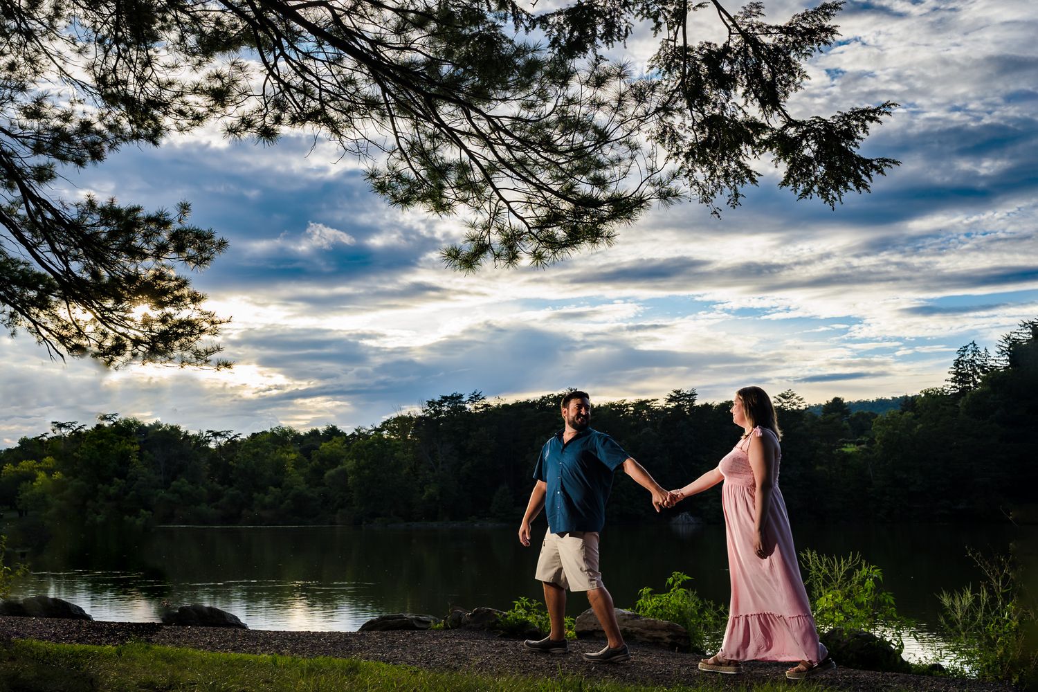 A couple walks along a lakeside path under dramatic clouds and overhanging trees.
