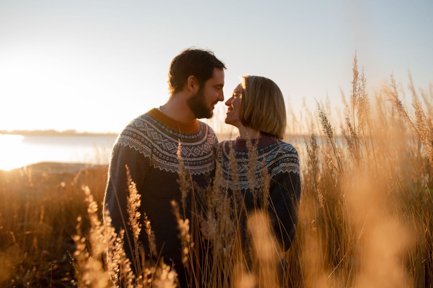 Couple in matching knit sweaters share an intimate moment among tall grass at sunset.