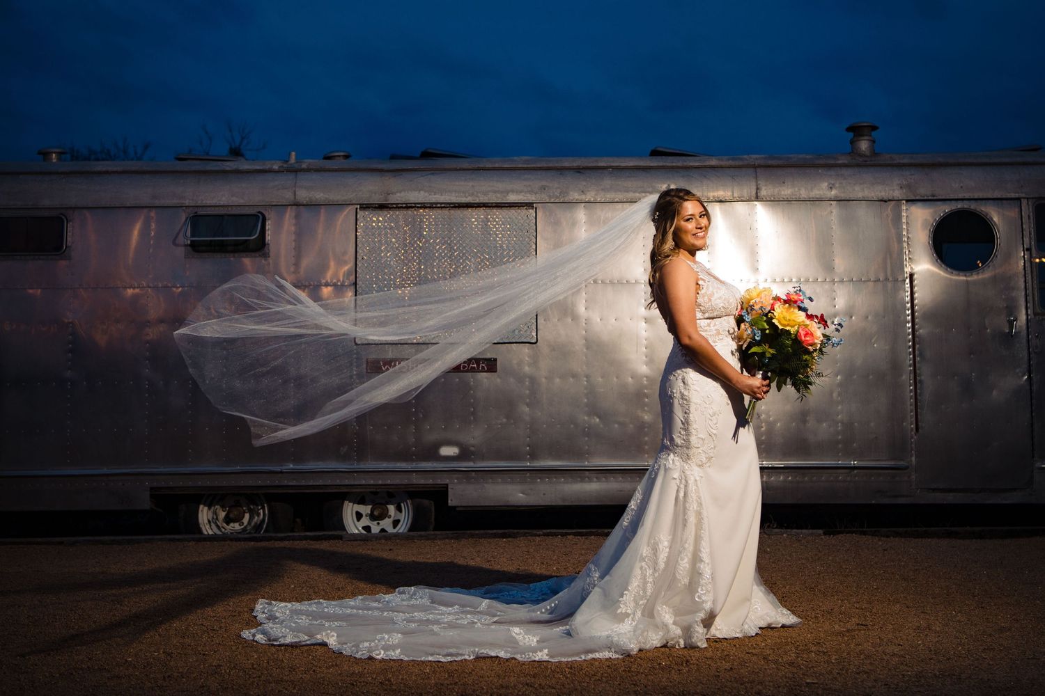 Bride in lace gown with flowing veil poses against vintage silver airstream trailer at dusk.