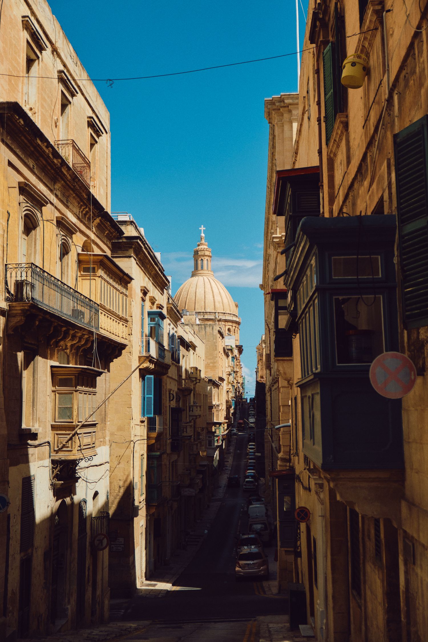 Narrow cobblestone street in Malta with historic buildings and church dome visible at the end under blue sky.