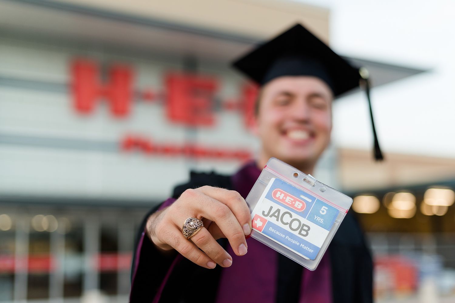 Graduate showing name tag at H-E-B store wearing cap and gown.