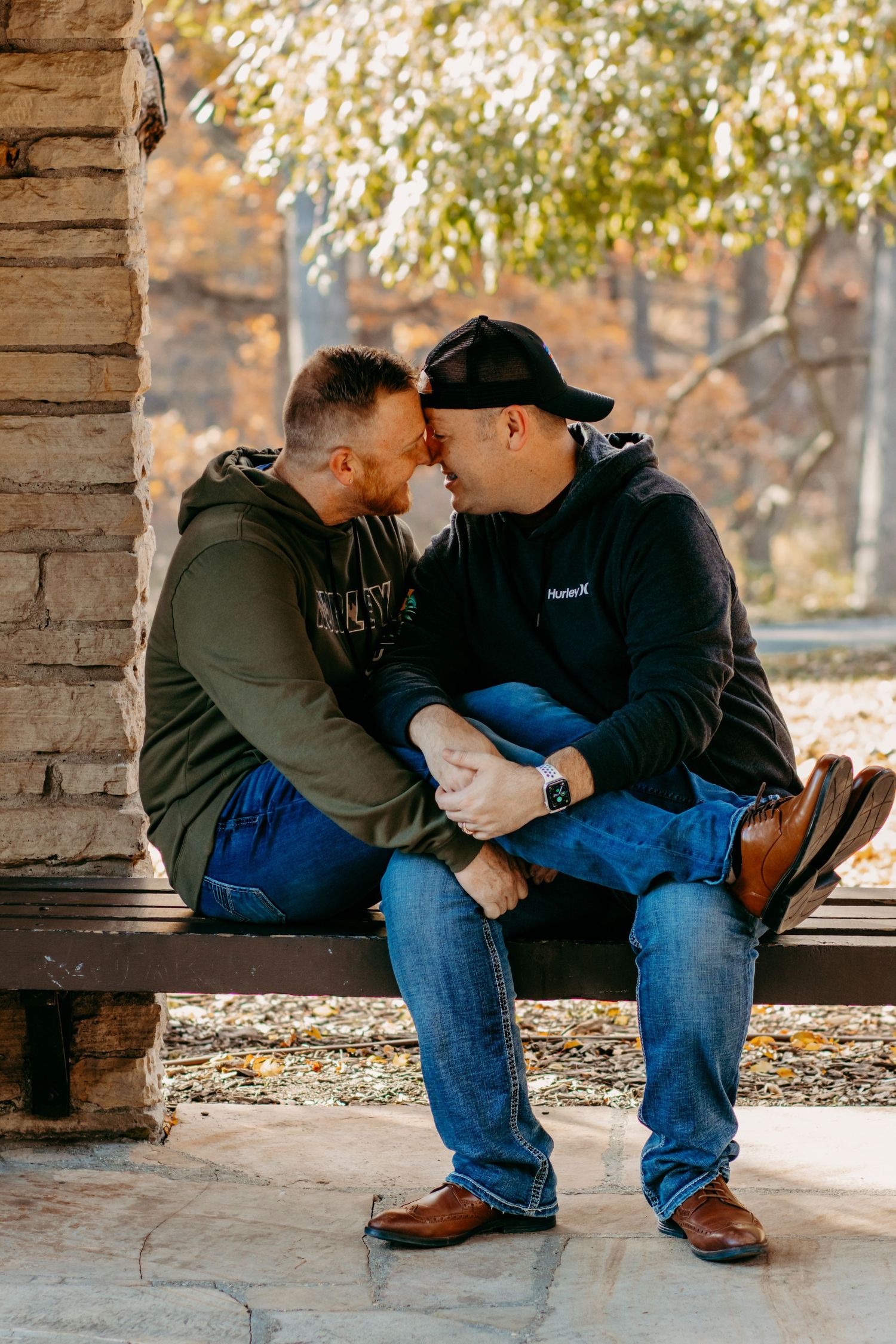 Couple shares intimate moment while sitting on stone bench surrounded by autumn trees.