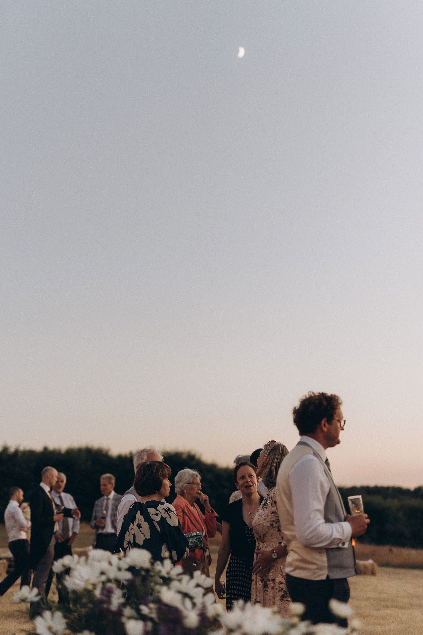 Wedding guests gather outdoors at dusk with the moon visible in the pale sky.
