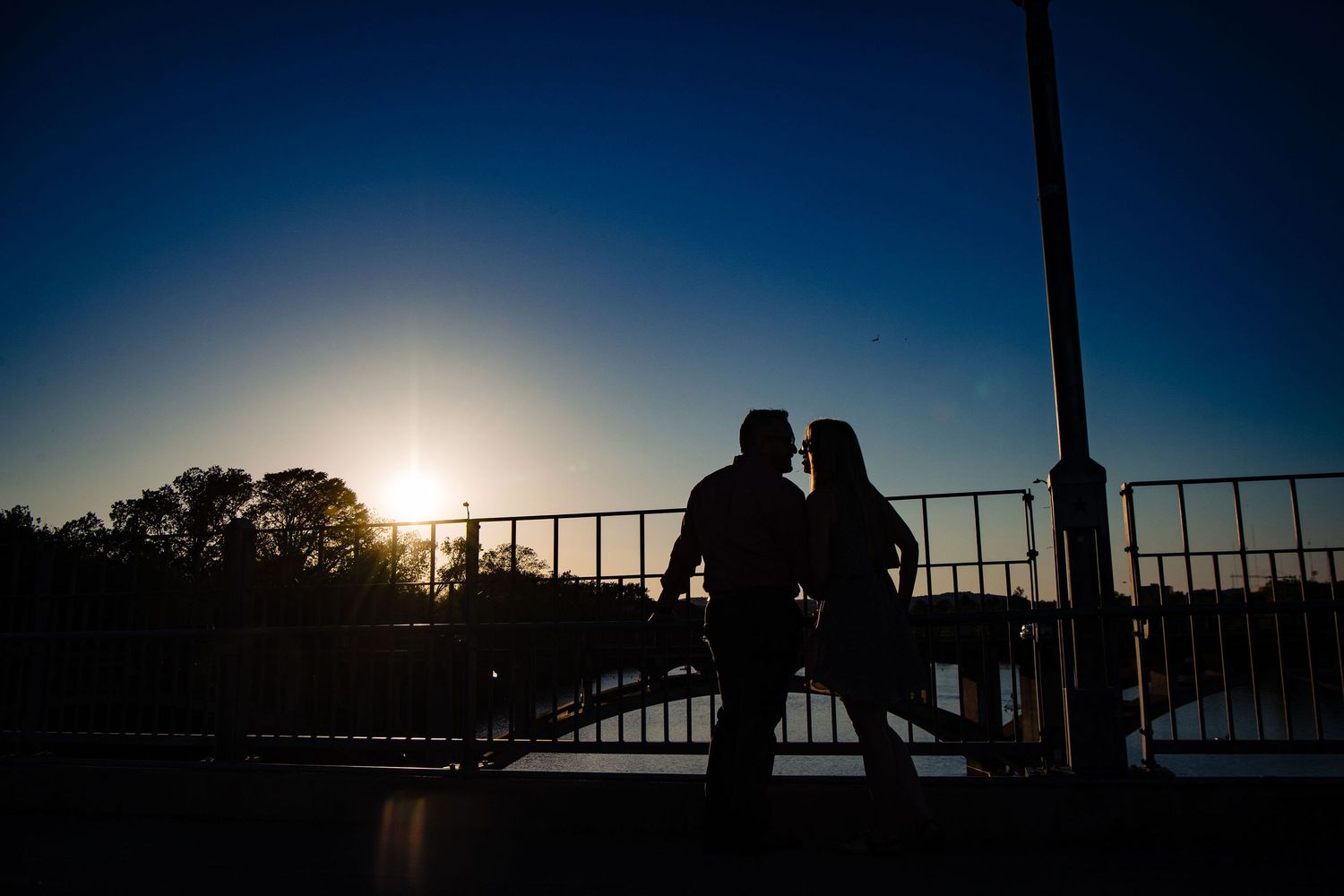 A silhouetted couple shares a romantic moment on a bridge during sunset.
