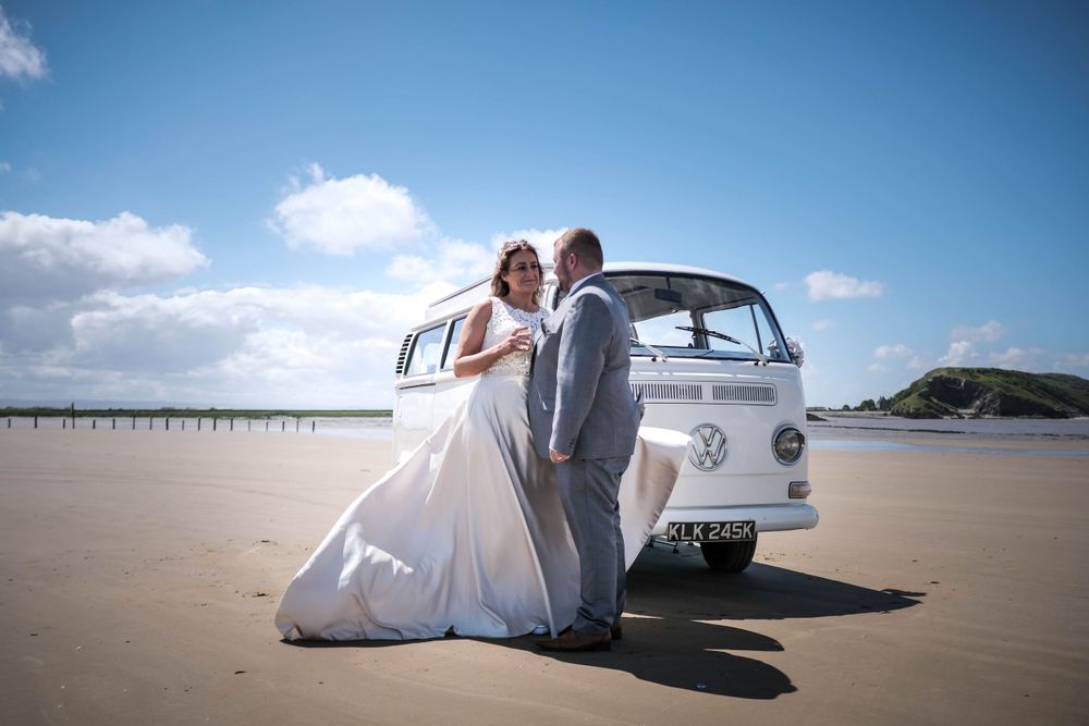 Newlyweds pose with classic white VW camper van on a scenic beach.