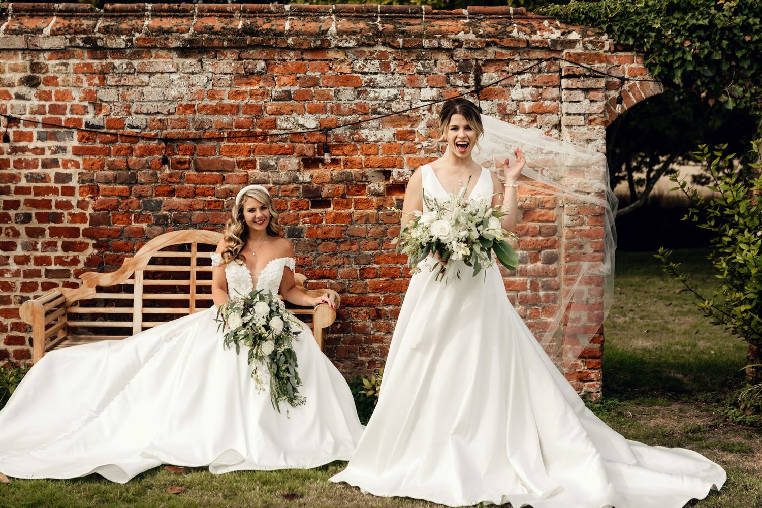 Two brides in elegant white wedding gowns with floral bouquets pose together against a rustic brick wall outdoors.