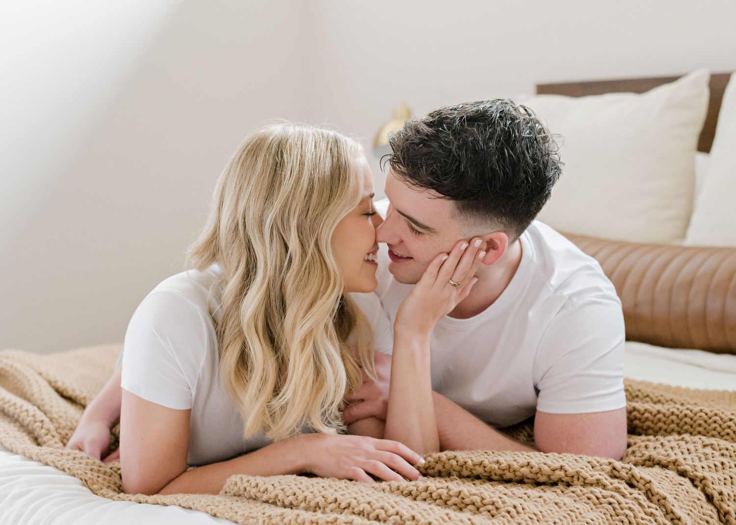 couple laying together on their bed, her hand up on his face, smiling and leaning in for a kiss