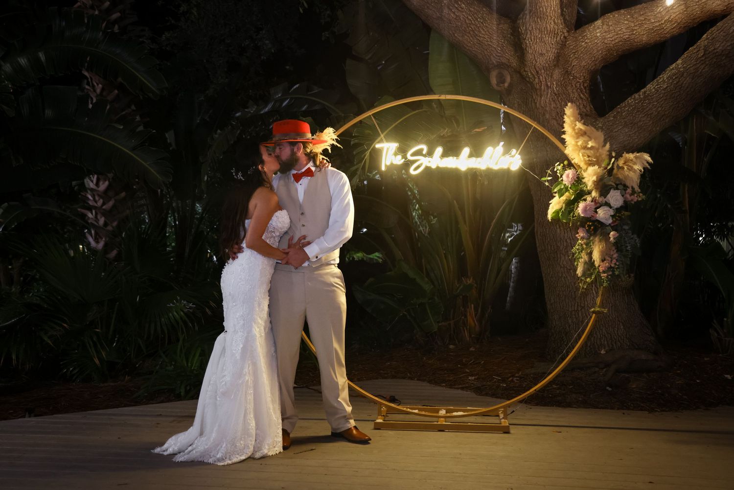 Romantic nighttime wedding photo with illuminated neon sign and circular backdrop in a garden setting.