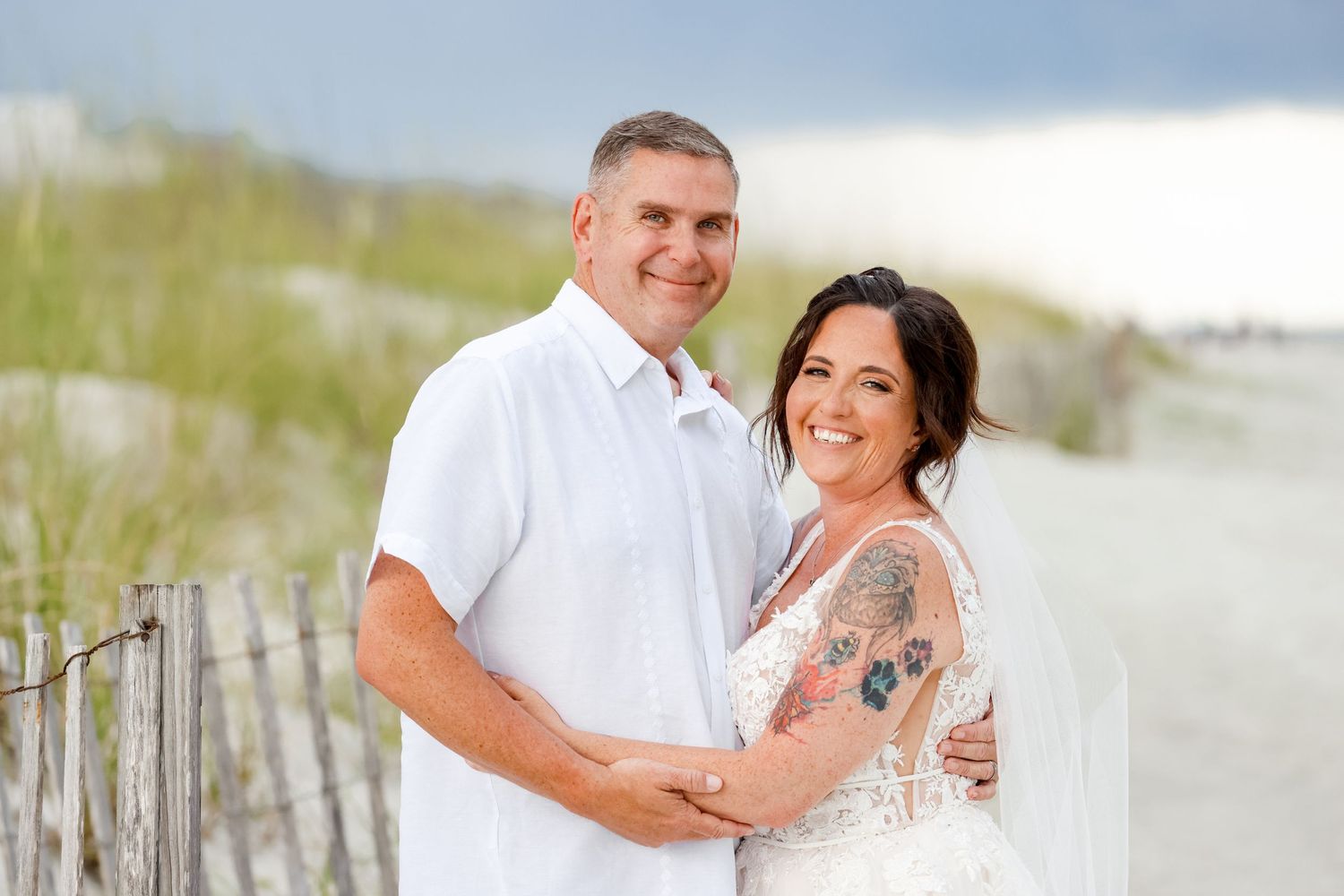 Couple embraces on beach boardwalk with sand dunes and ocean in background.