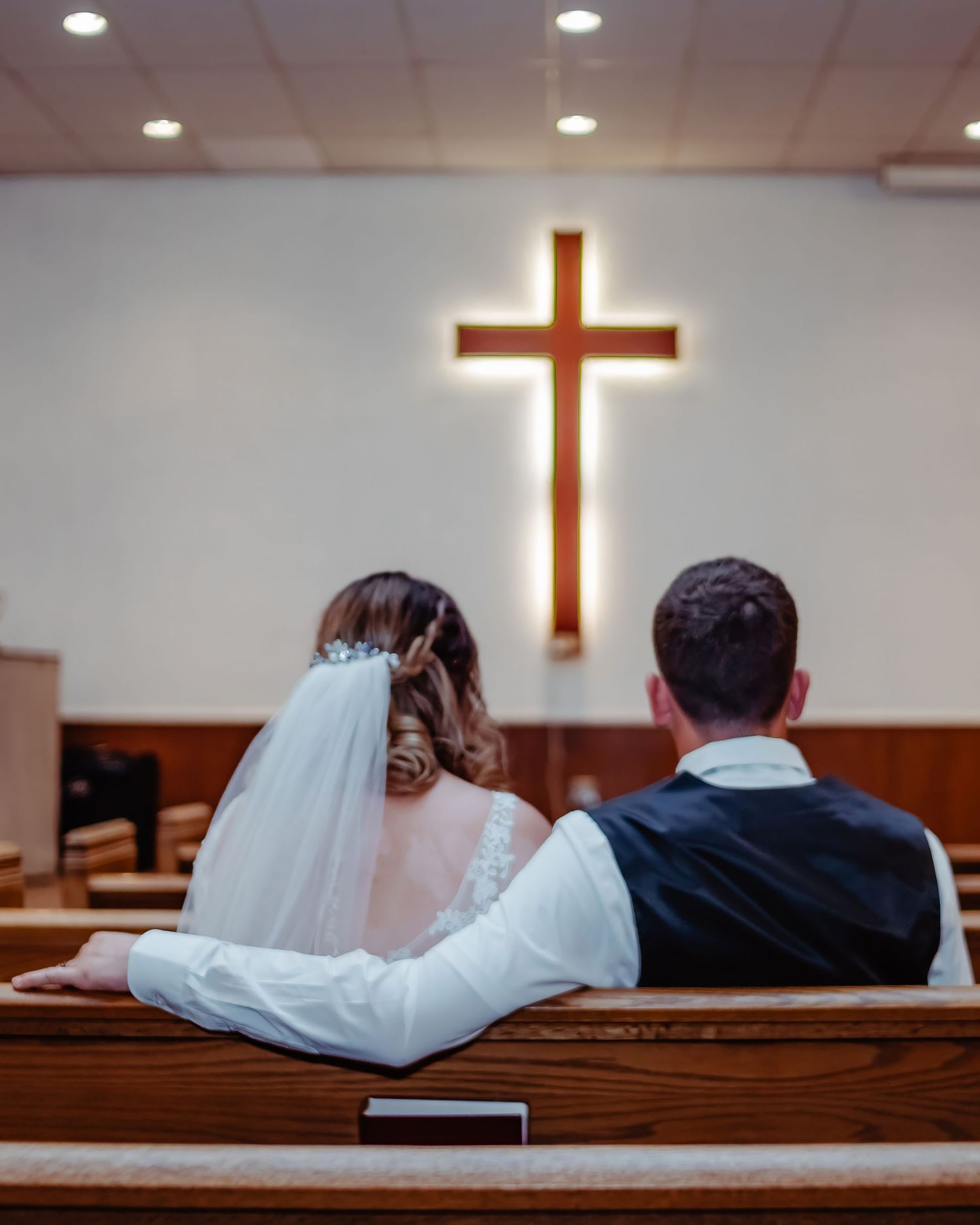 A couple sits together on a wooden church pew with an illuminated cross glowing on the wall behind them.