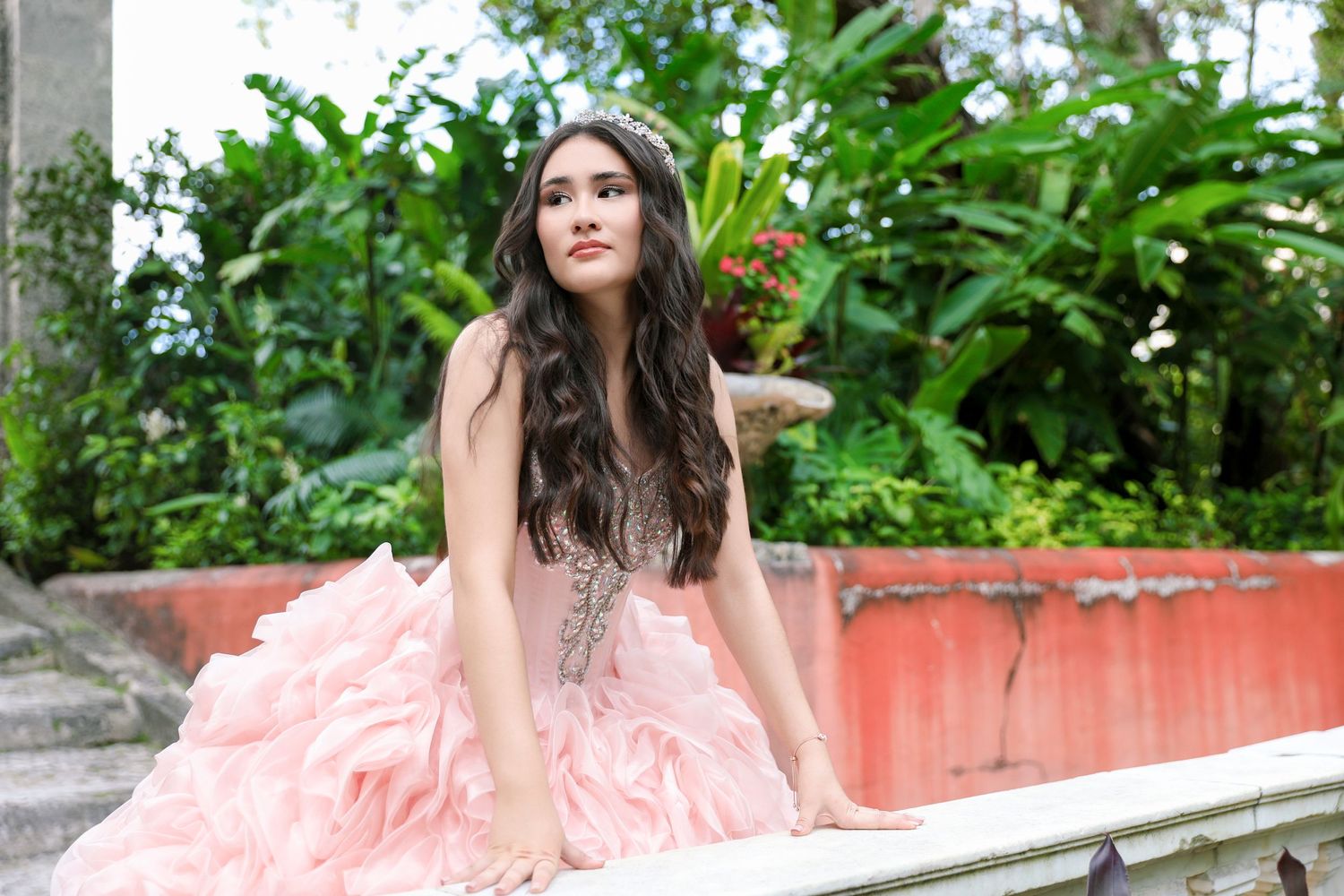 Elegant quinceañera portrait in coral pink ruffled ballgown amid tropical foliage.