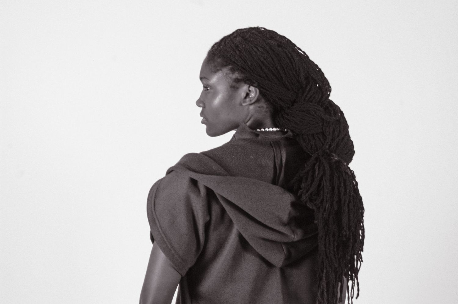 Black and white portrait of a woman with long dreadlocks wearing a dark hoodie, shot from behind.
