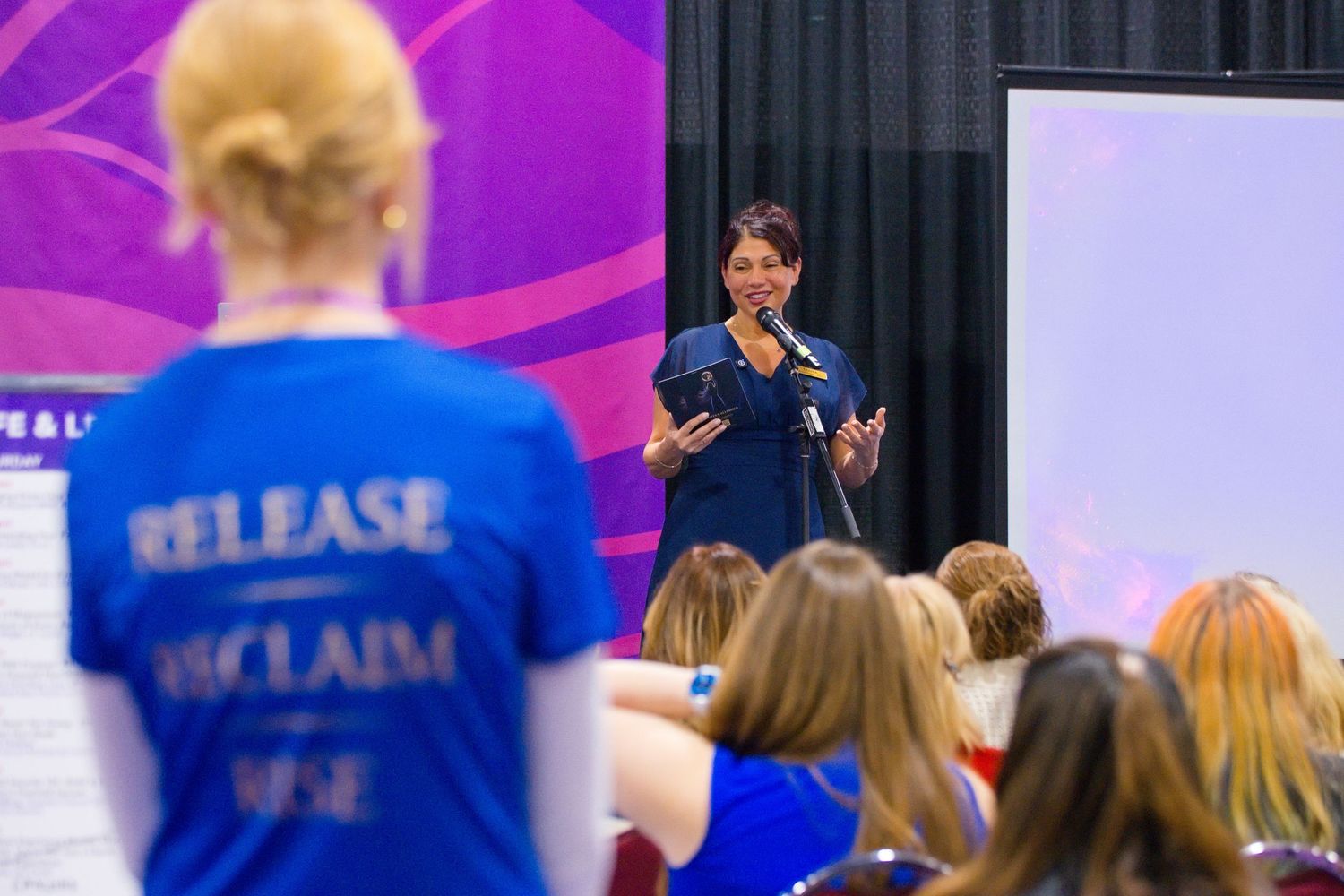 Woman in a blue shirt speaks into a microphone to a seated audience, while an attendee in a Release Reclaim Rise shirt looks on.
