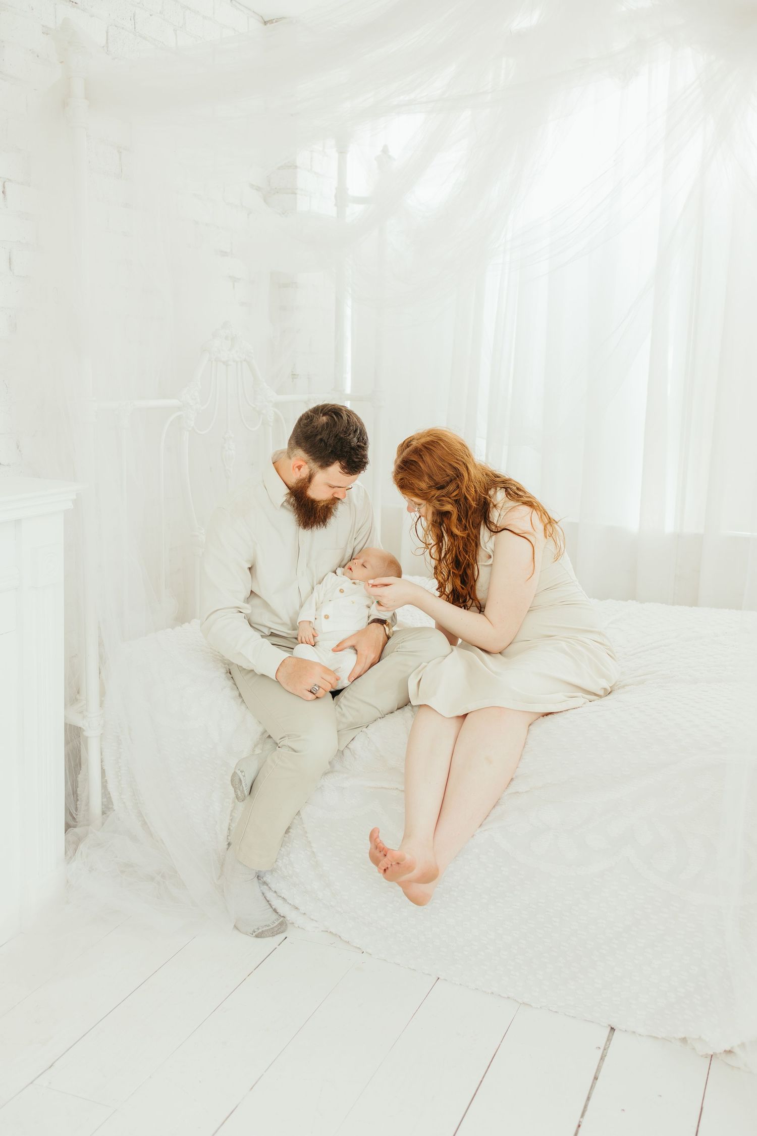 A bearded father and redhead mother sit on a white bed together, lovingly attending to their newborn baby.