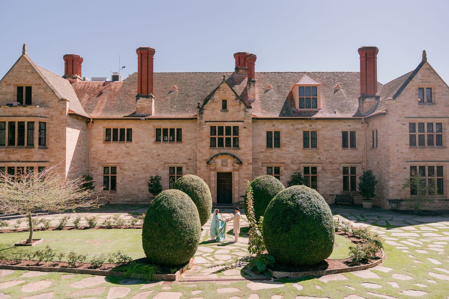Historic stone manor house with tall brick chimneys and mullioned windows overlooking a sunlit courtyard garden with rounded topiary shrubs.