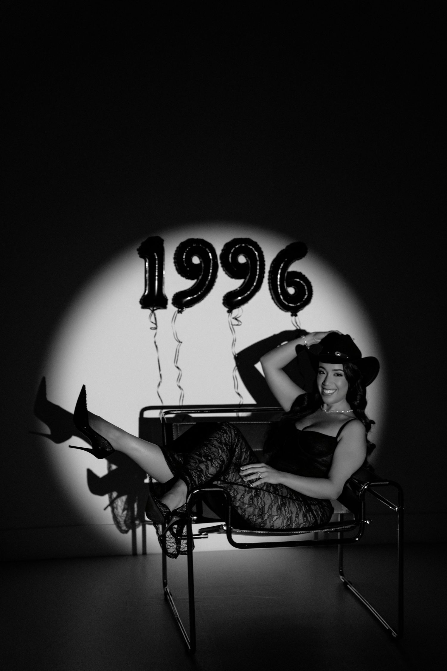 Black and white photo of a smiling woman in a cowboy hat posing in a chair with 1996 balloons in a spotlight.