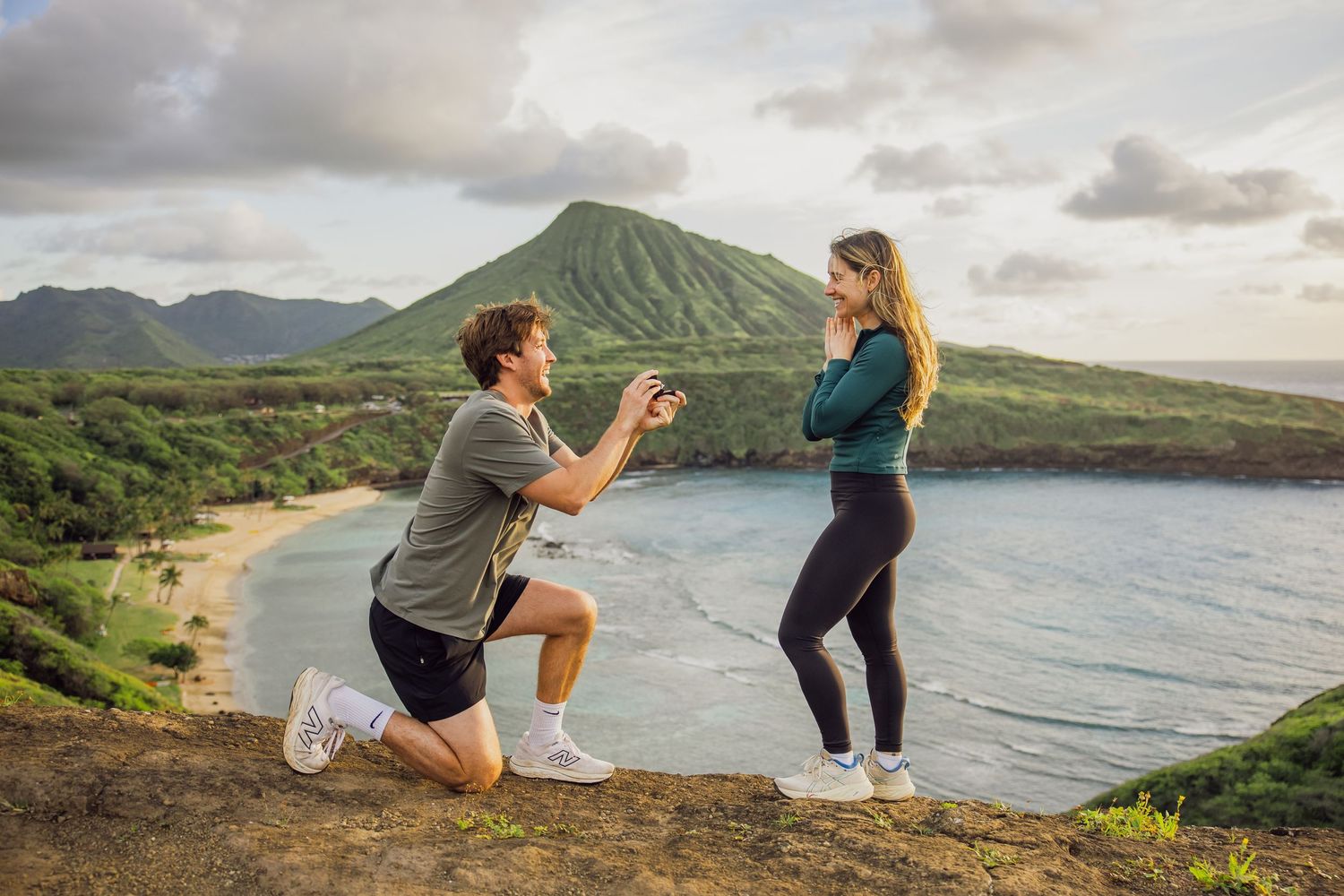 Man proposes to woman on scenic Hawaiian cliff overlooking a beach with lush green mountains in the background.