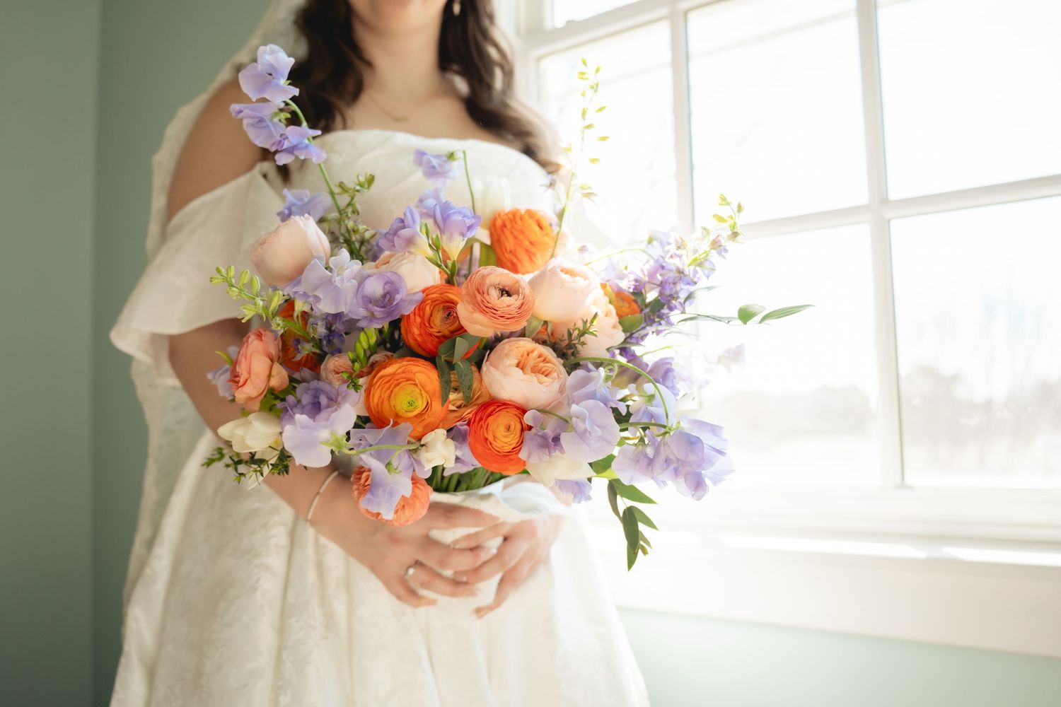 Bride in white off-shoulder gown holding vibrant bouquet of orange ranunculus and purple wildflowers near window.