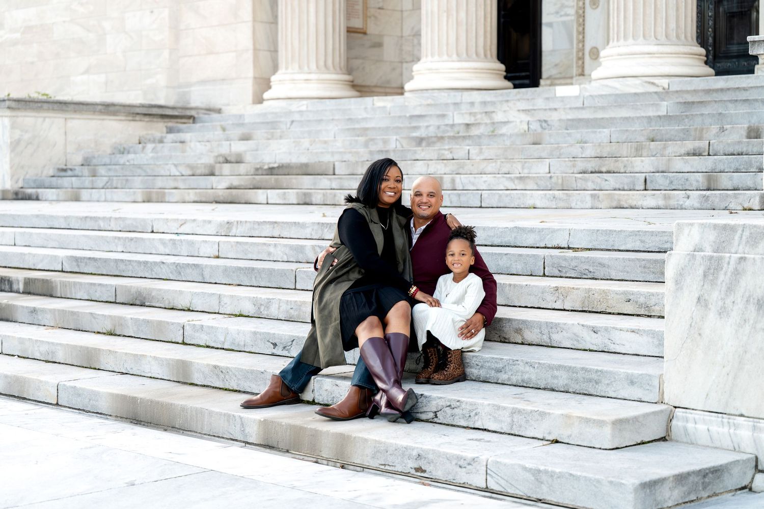 Family of three sitting on marble steps of a grand building with white columns, smiling together outdoors.