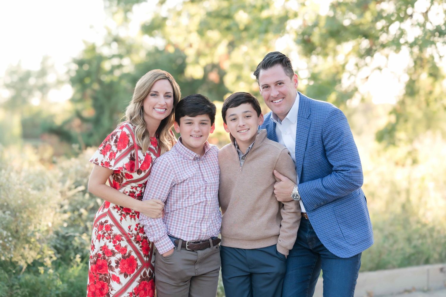 A smiling family of four poses outdoors in warm evening light, wearing smart casual attire in a natural garden setting.