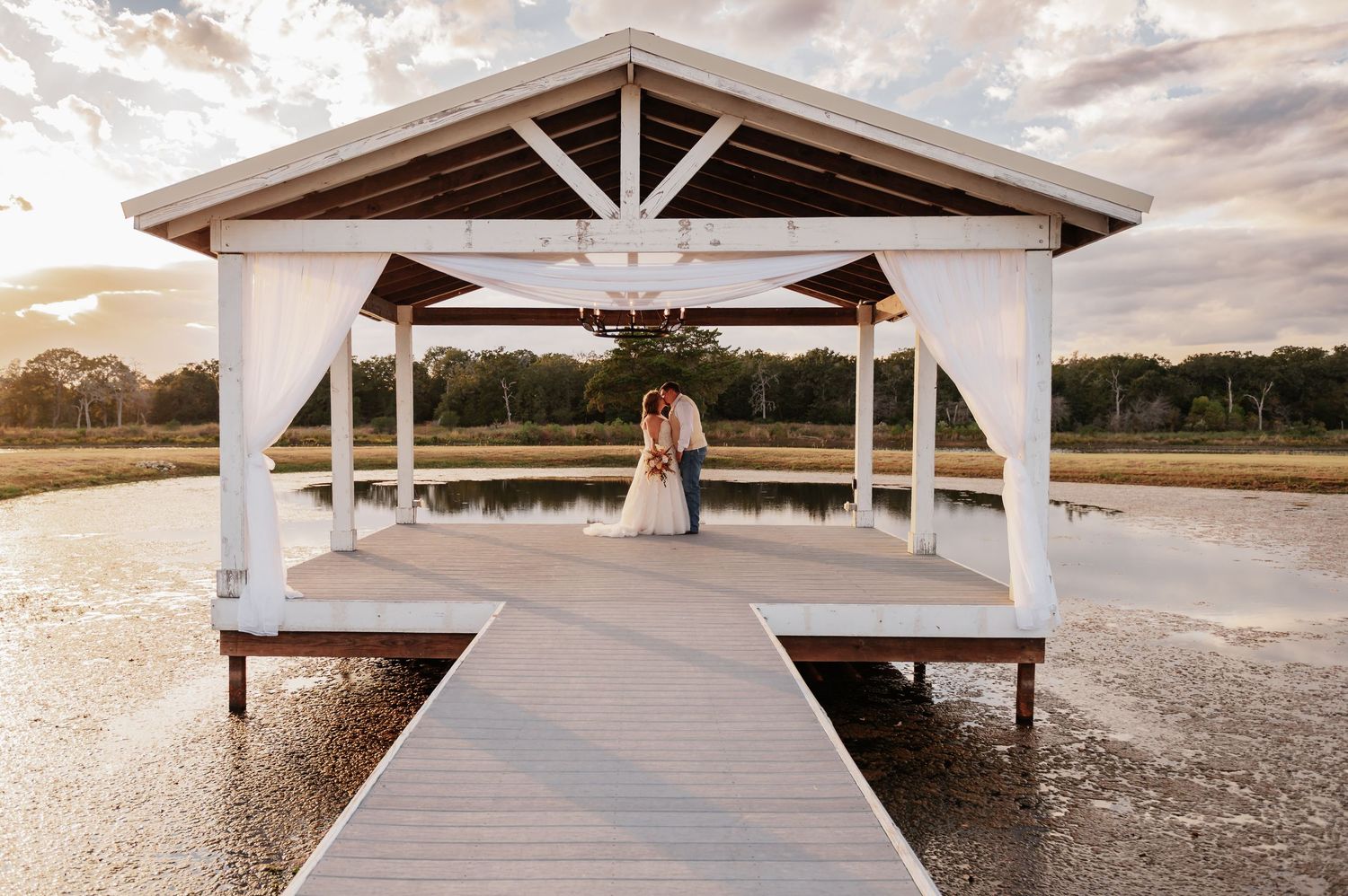 Bride and groom exchange vows at rustic indoor wedding ceremony with white chairs and floral arrangements.