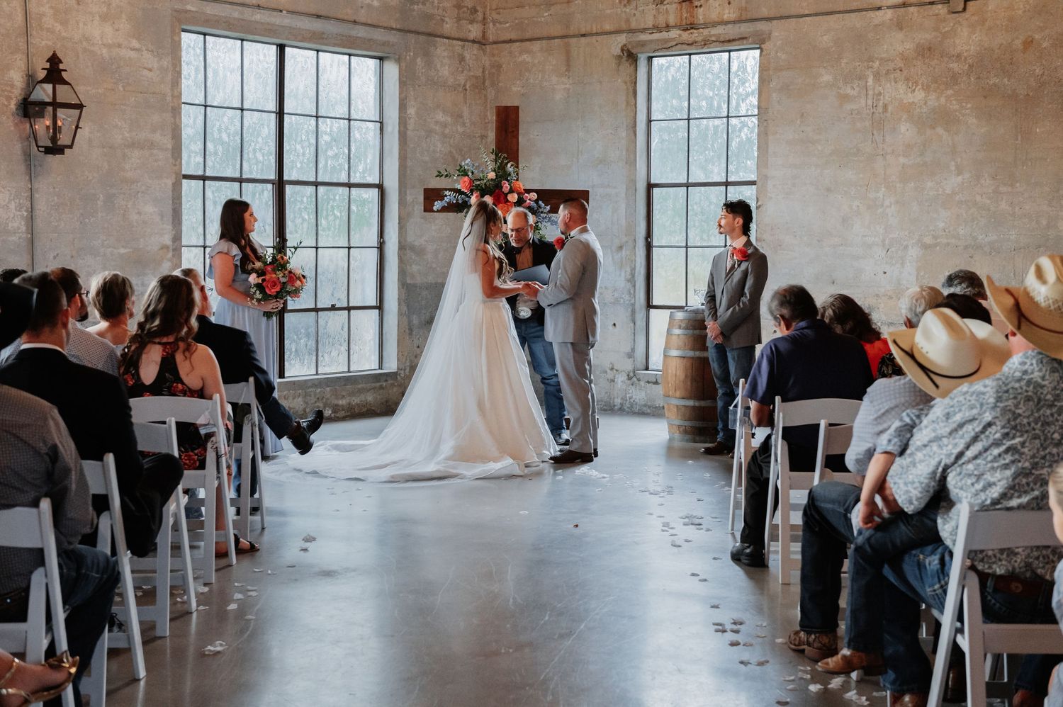 Bride and groom exchange vows at rustic indoor wedding ceremony with white chairs and floral arrangements.