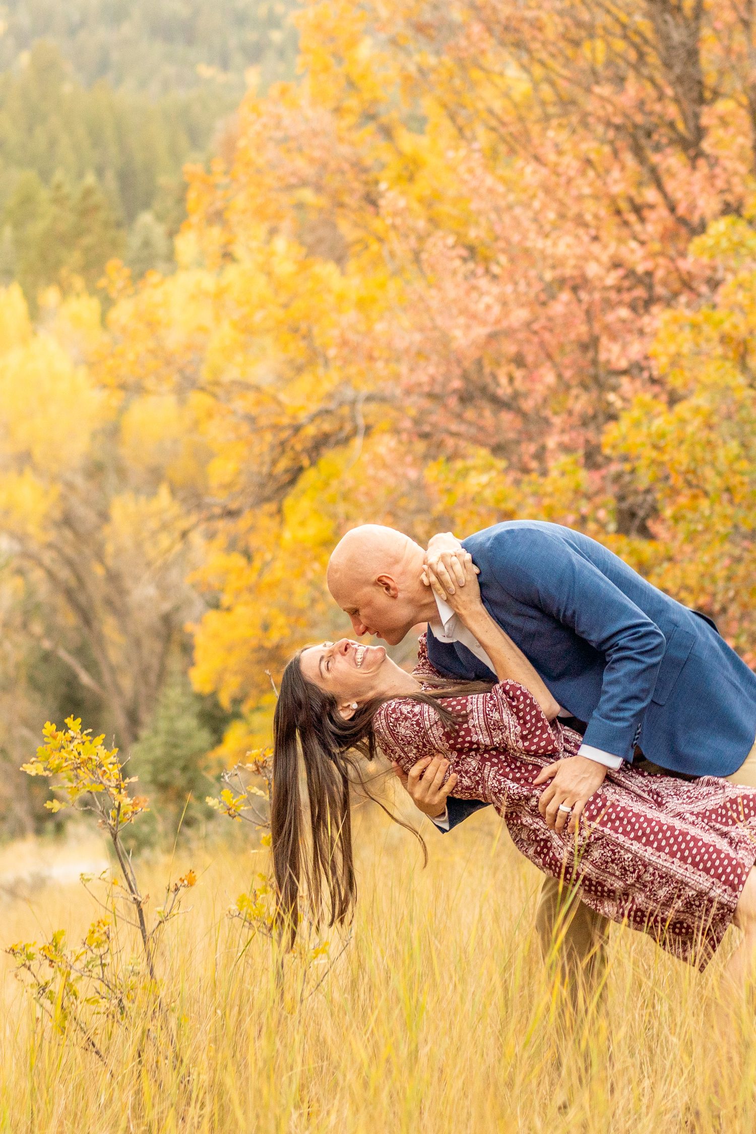 Romantic couple embraces in tall golden grass with vibrant autumn foliage creating a warm seasonal backdrop.
