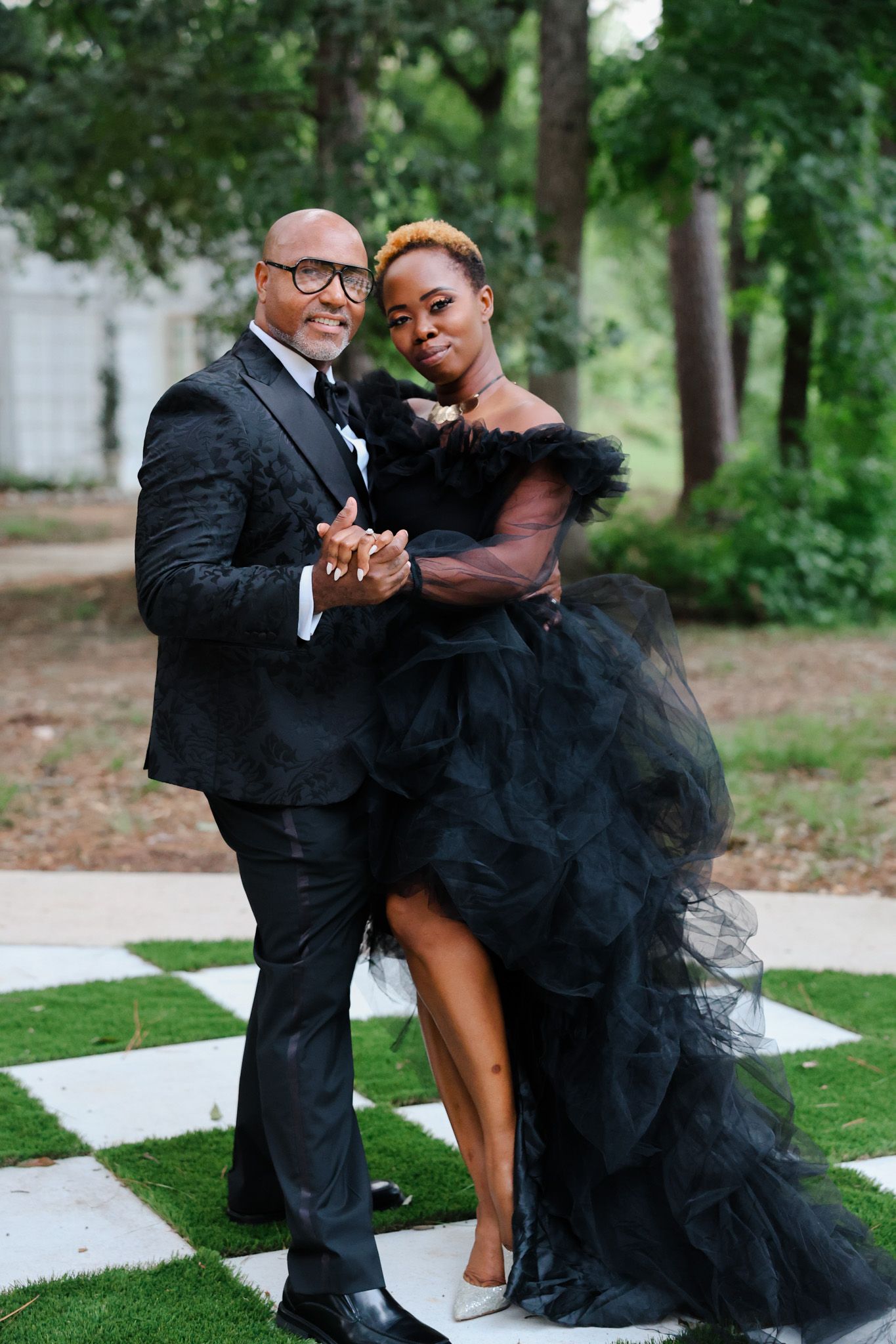 A couple in formal attire dance on a checkered outdoor patio surrounded by trees.