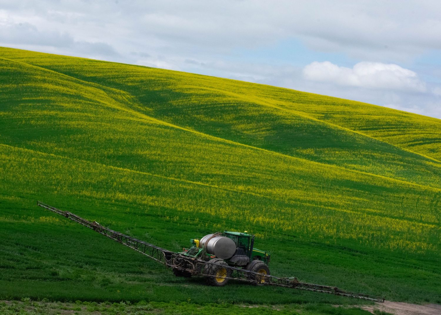 John Deere sprayer working across vibrant green and yellow rolling crop fields under cloudy sky.