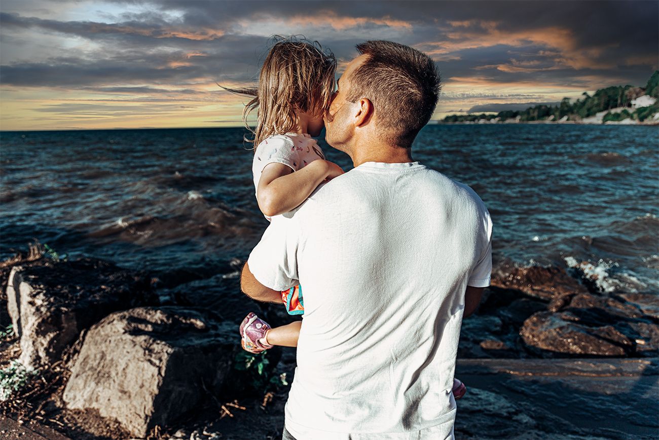 A white-shirted figure holds a child while standing on rocky coastal cliffs at sunset against dramatic ocean waves.