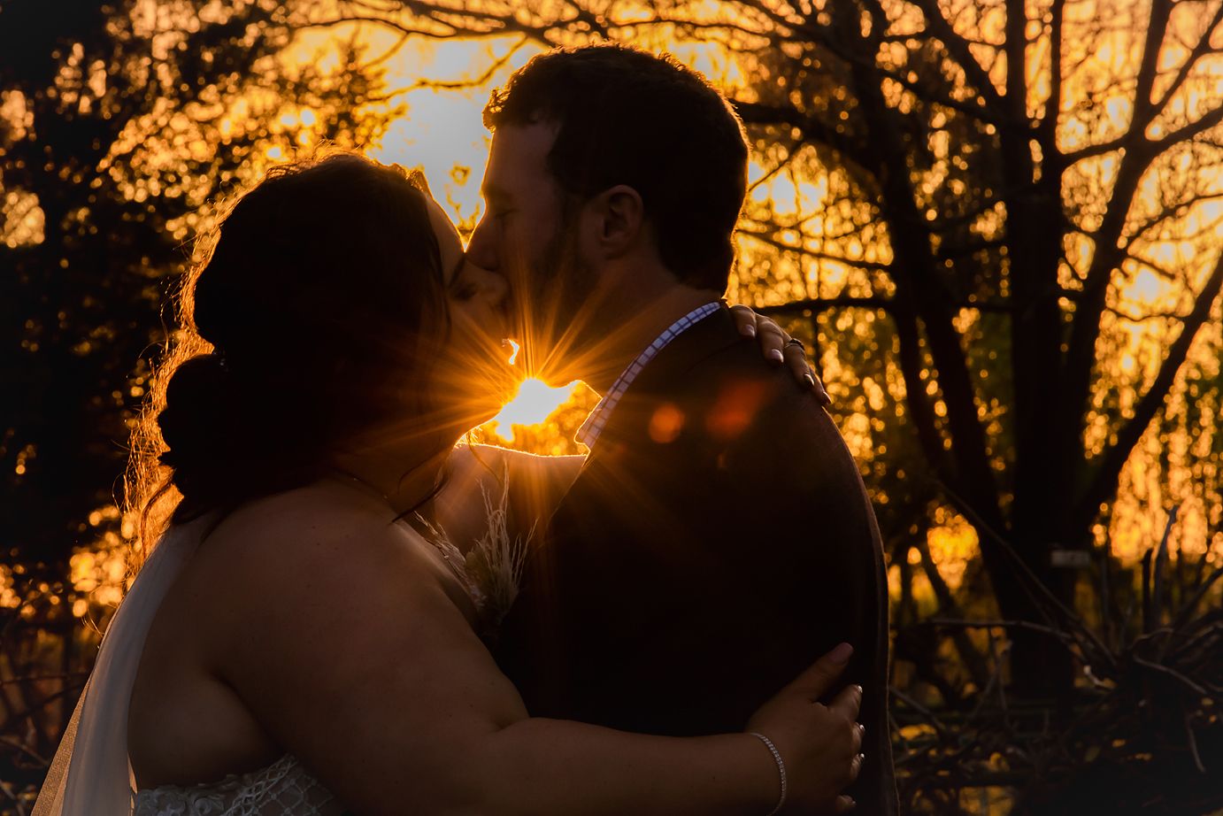 A silhouetted couple embraces during golden hour sunset with sunlight peeking through the trees behind them.