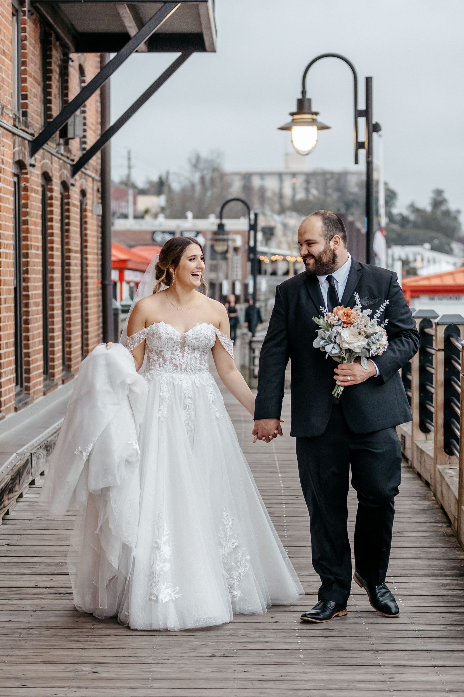 A newlywed couple walking together on a rustic wooden boardwalk holding hands as the bride carries her dress train.