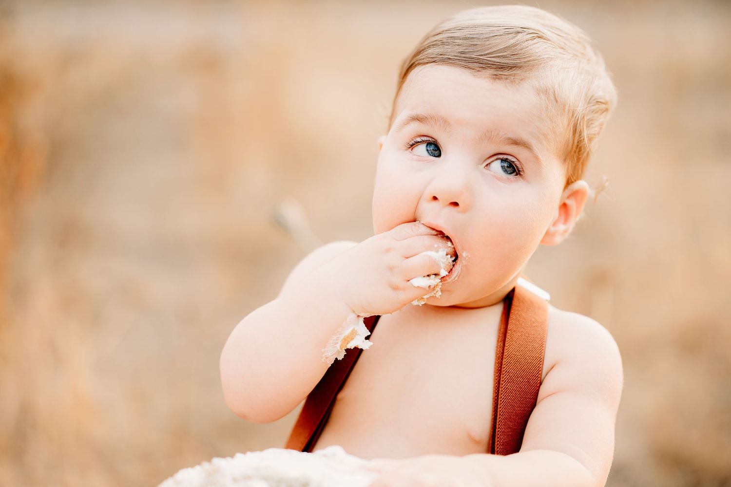 A toddler in brown suspenders looks upward with a curious expression while putting fingers in their mouth during first birthday photos.