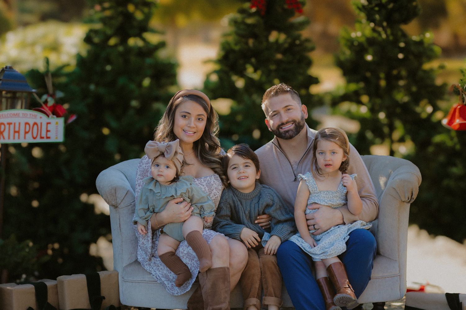 Family of six sits together on beige couch surrounded by Christmas trees and North Pole decor.