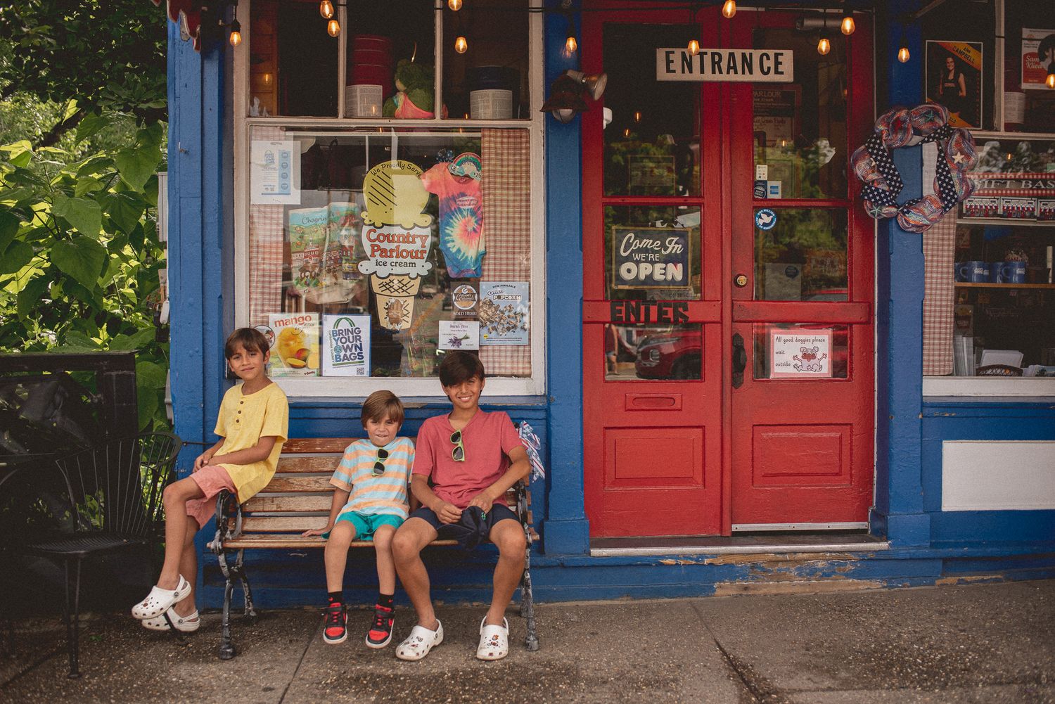People sitting on steps outside a colorful storefront with bright red doors and signage on a summer day.