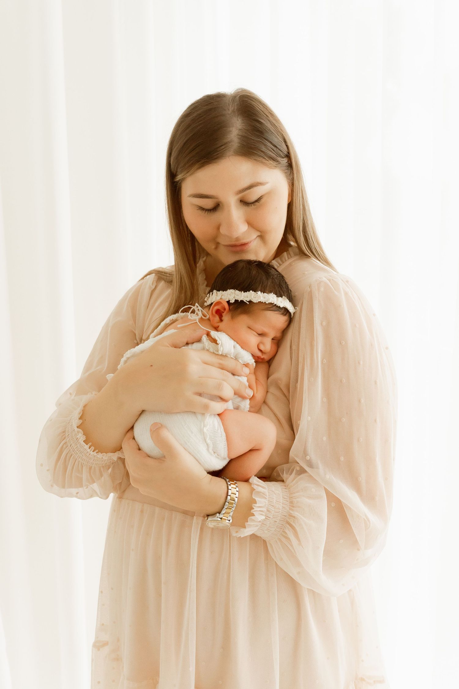 Mother in flowing white dress holding sleeping newborn baby against white curtain background.