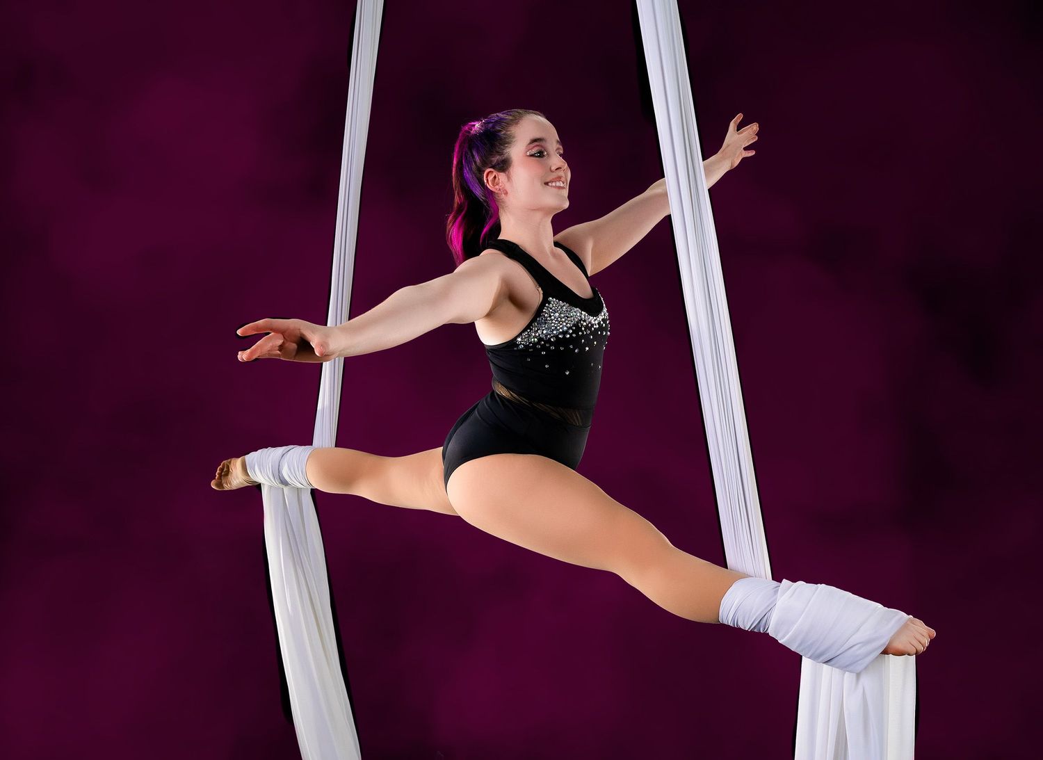 Aerial performer in splits pose on white silk ribbons against black background.
