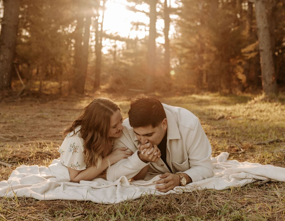 A couple lies together on a white blanket in a warm sunlit forest during golden hour.