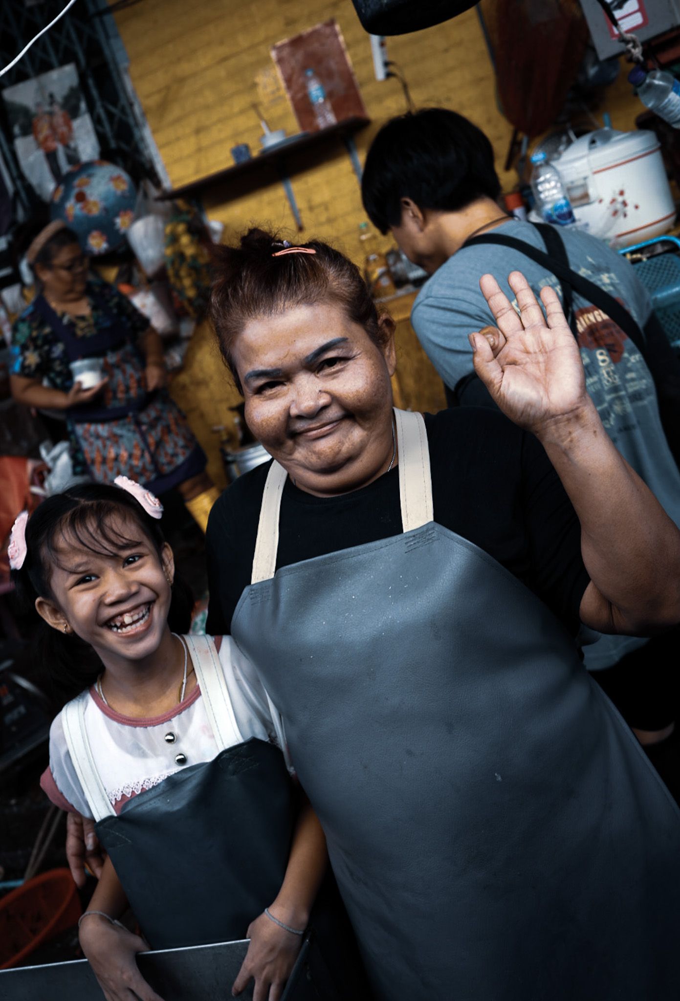 A smiling woman and young girl wearing gray aprons wave at the camera in a busy Asian street food market.