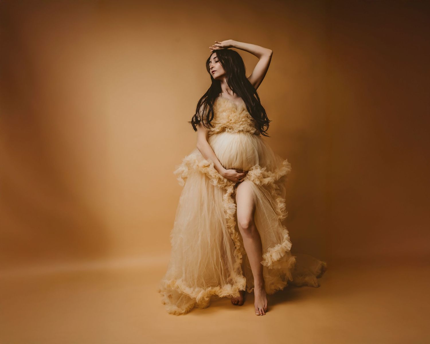 A person poses dramatically in a flowing beige tulle dress against a warm brown studio backdrop.