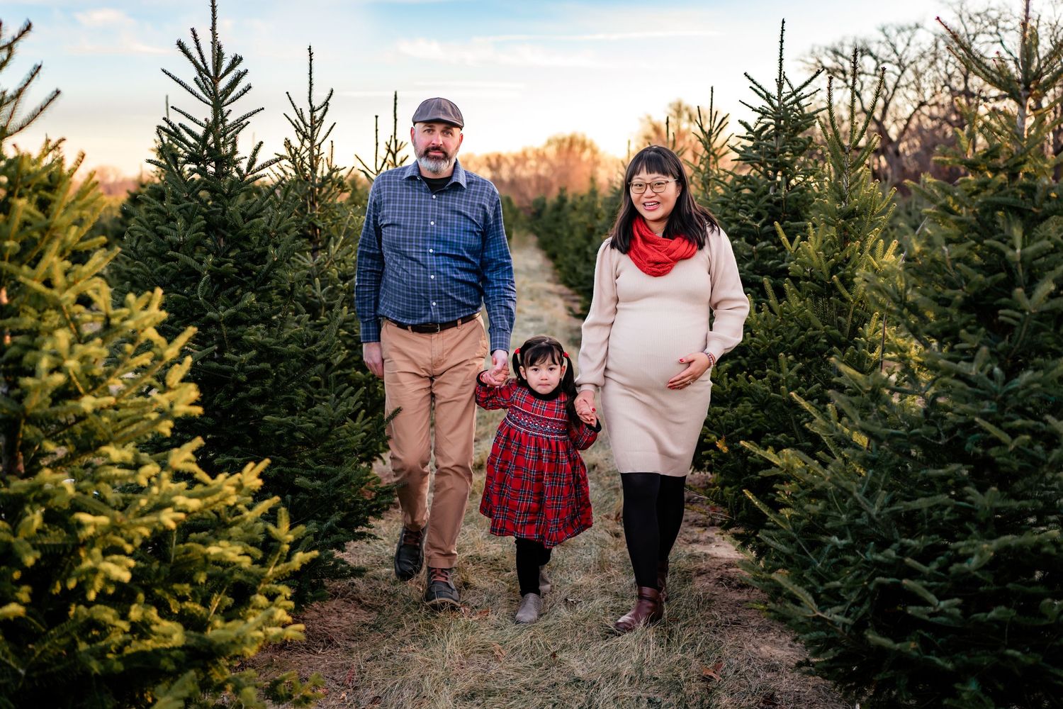 Three people walk together between rows of evergreen Christmas trees at sunset in a tree farm.