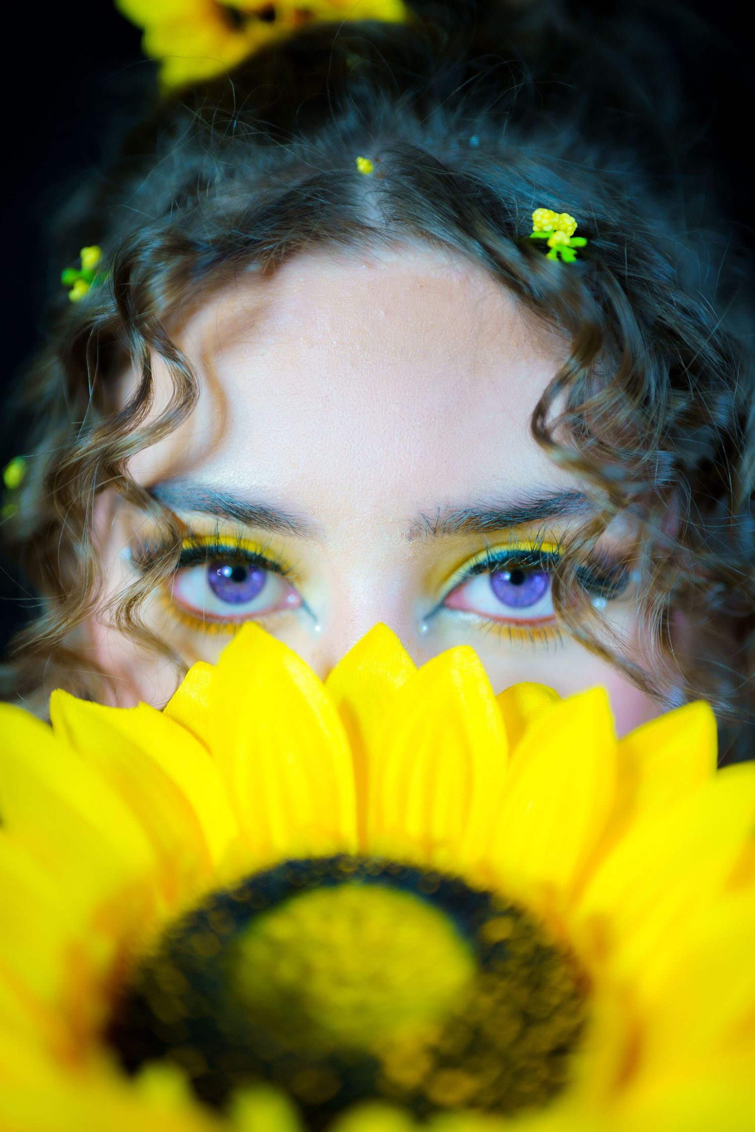Vibrant blue eyes peek over a bright yellow sunflower bloom with small green floral accents in curly dark hair.