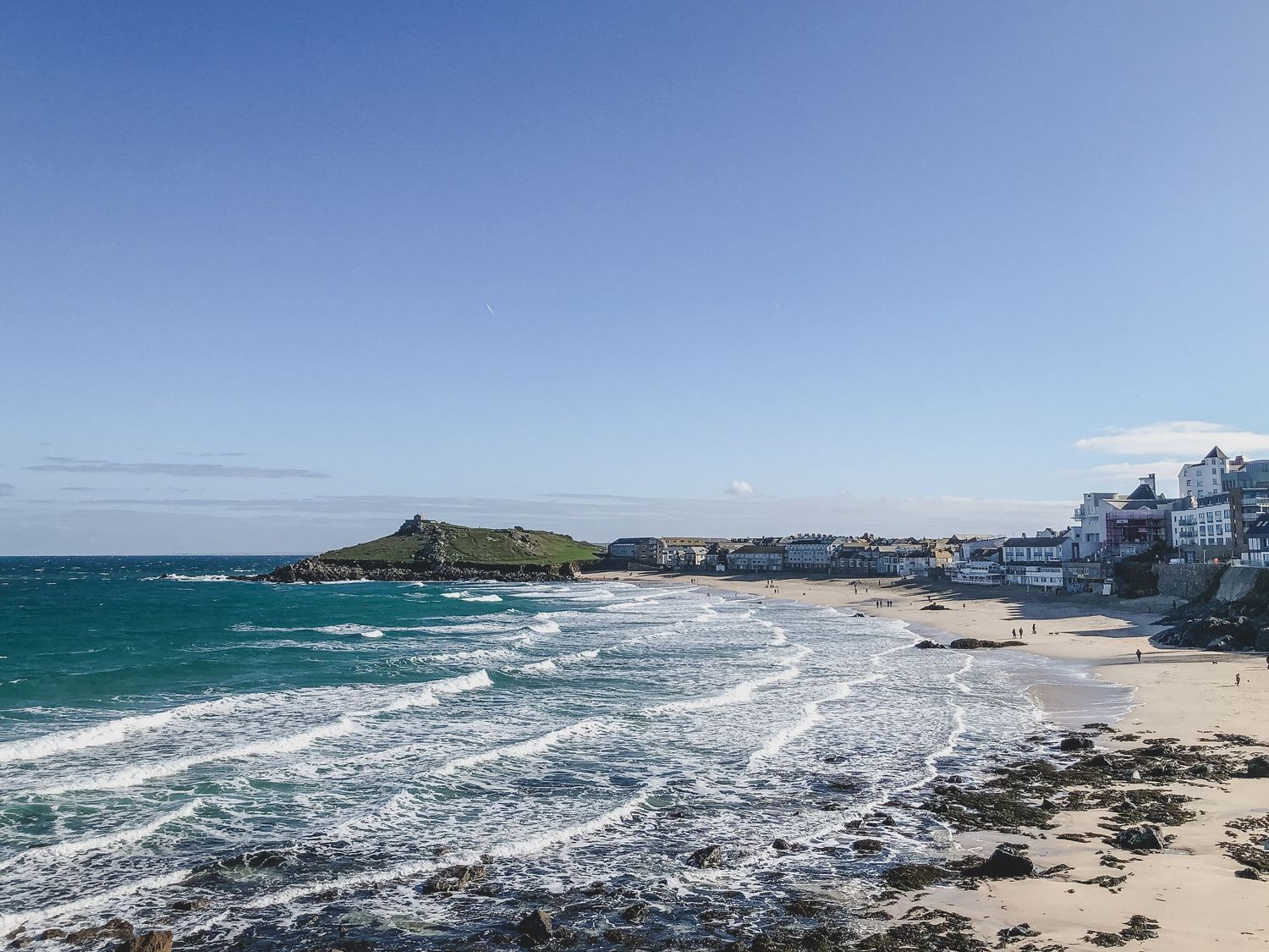Panoramic view of St Ives Bay in Cornwall showing turquoise waters, sandy beach and coastal buildings against blue sky.