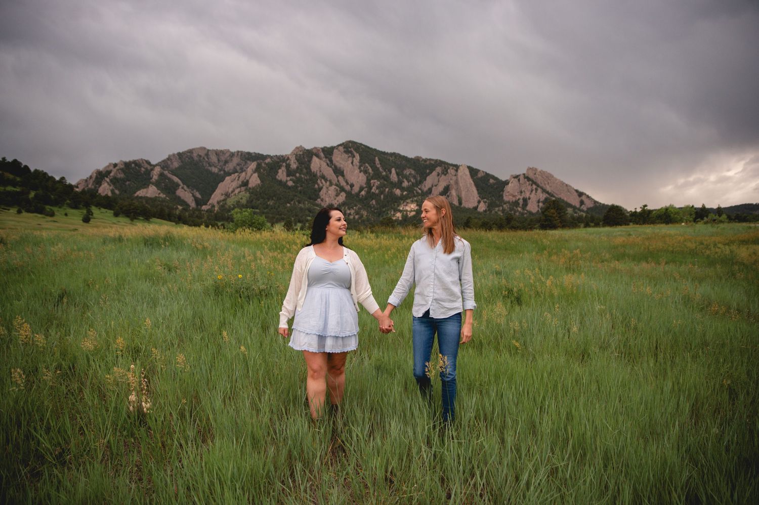 A couple walks together through a green mountain meadow with the majestic Flatirons rock formation visible in the distance.