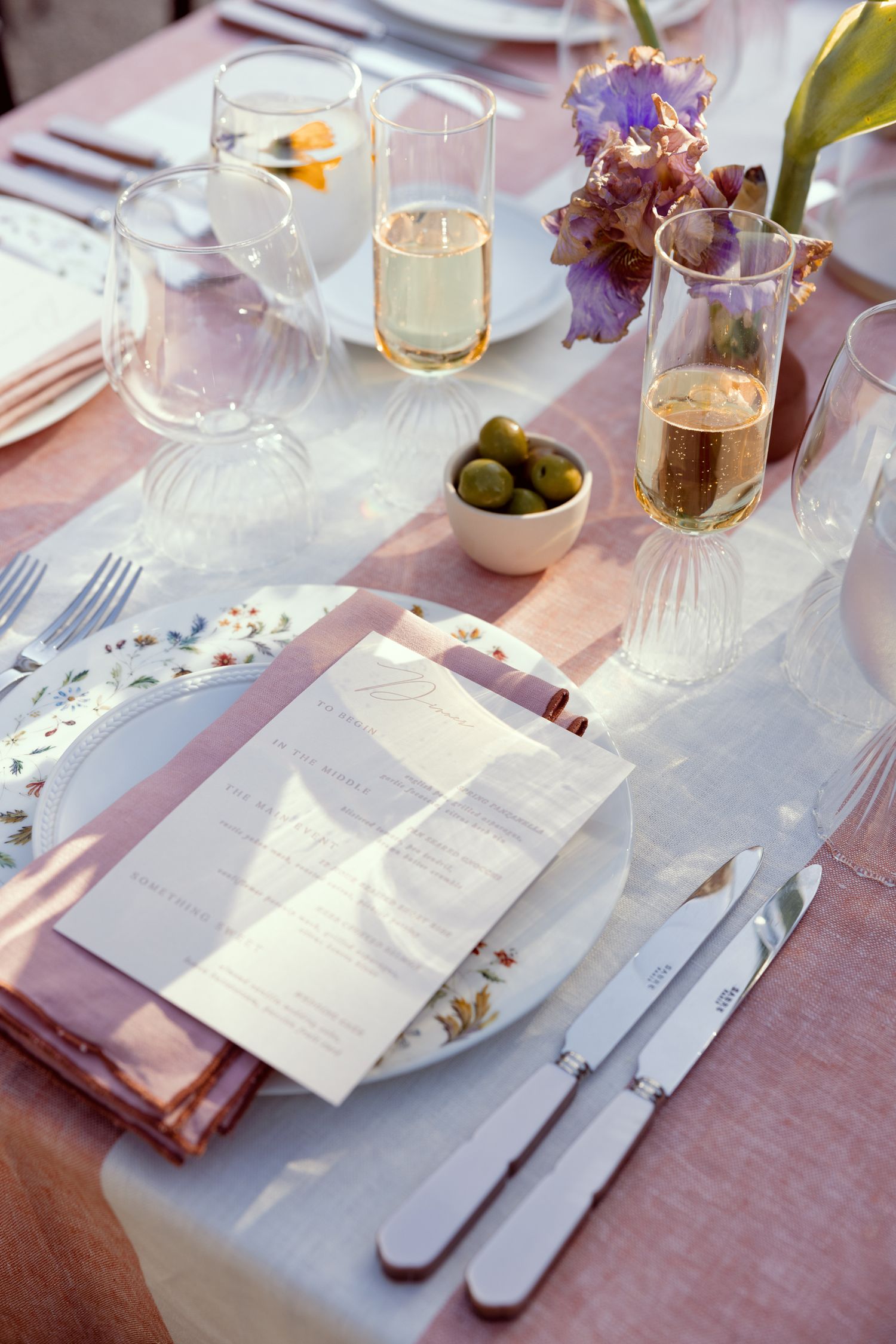 Elegant table setting with menu card, pink napkins, champagne glasses, olive bowl and purple flowers on white tablecloth.