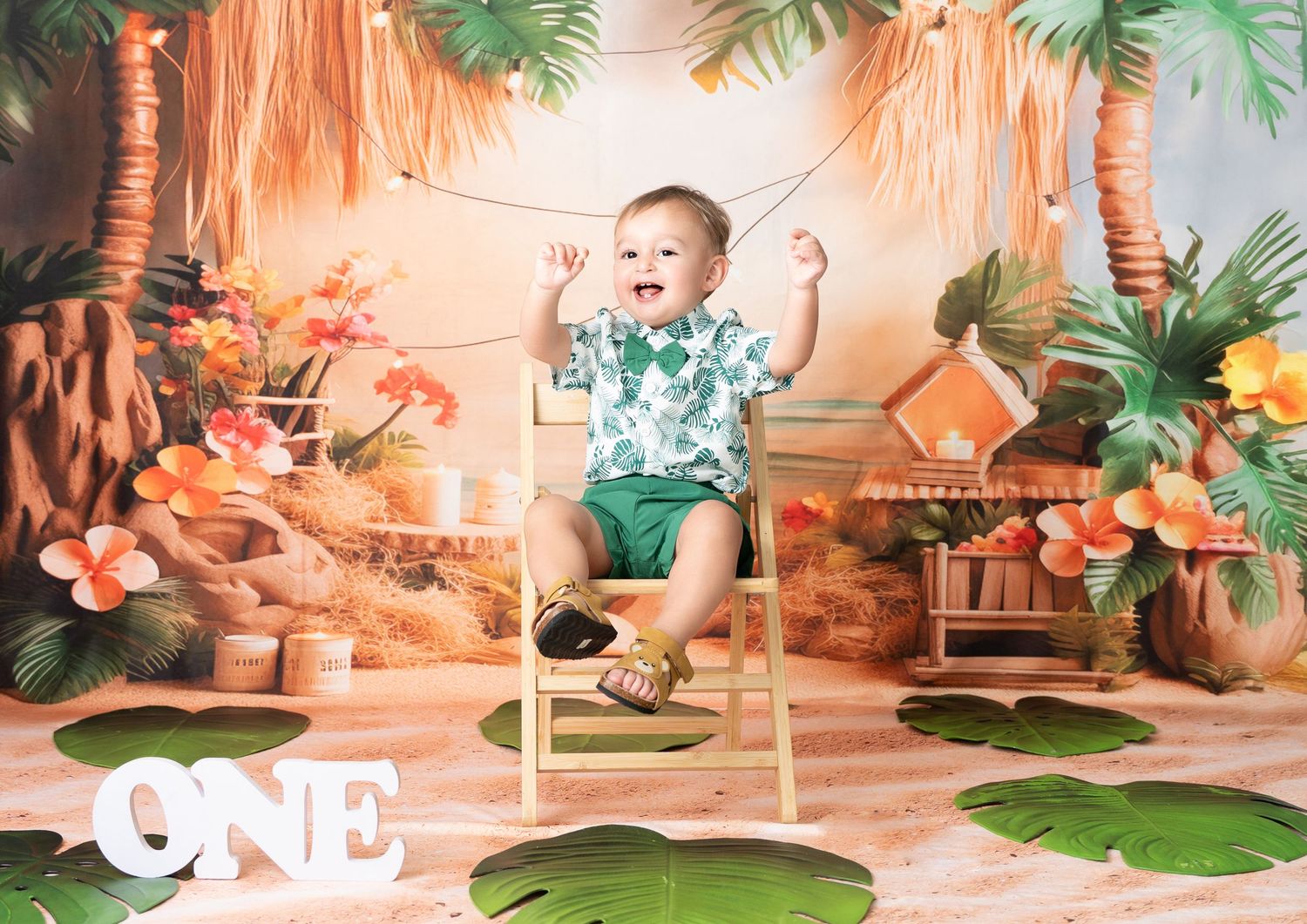 Baby sits among tropical palm decor, wearing a floral Hawaiian shirt and shorts set in green and white tones.